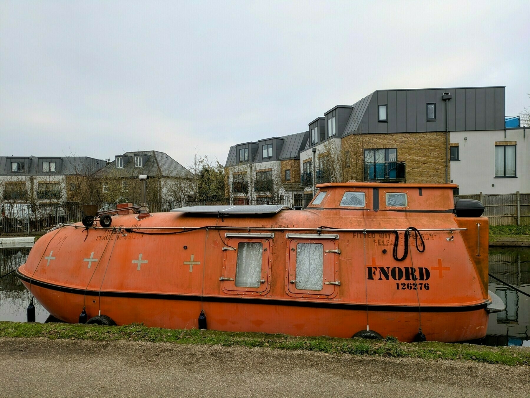 An orange lifeboat labeled FNORD is moored on a canal with modern townhouses in the background.