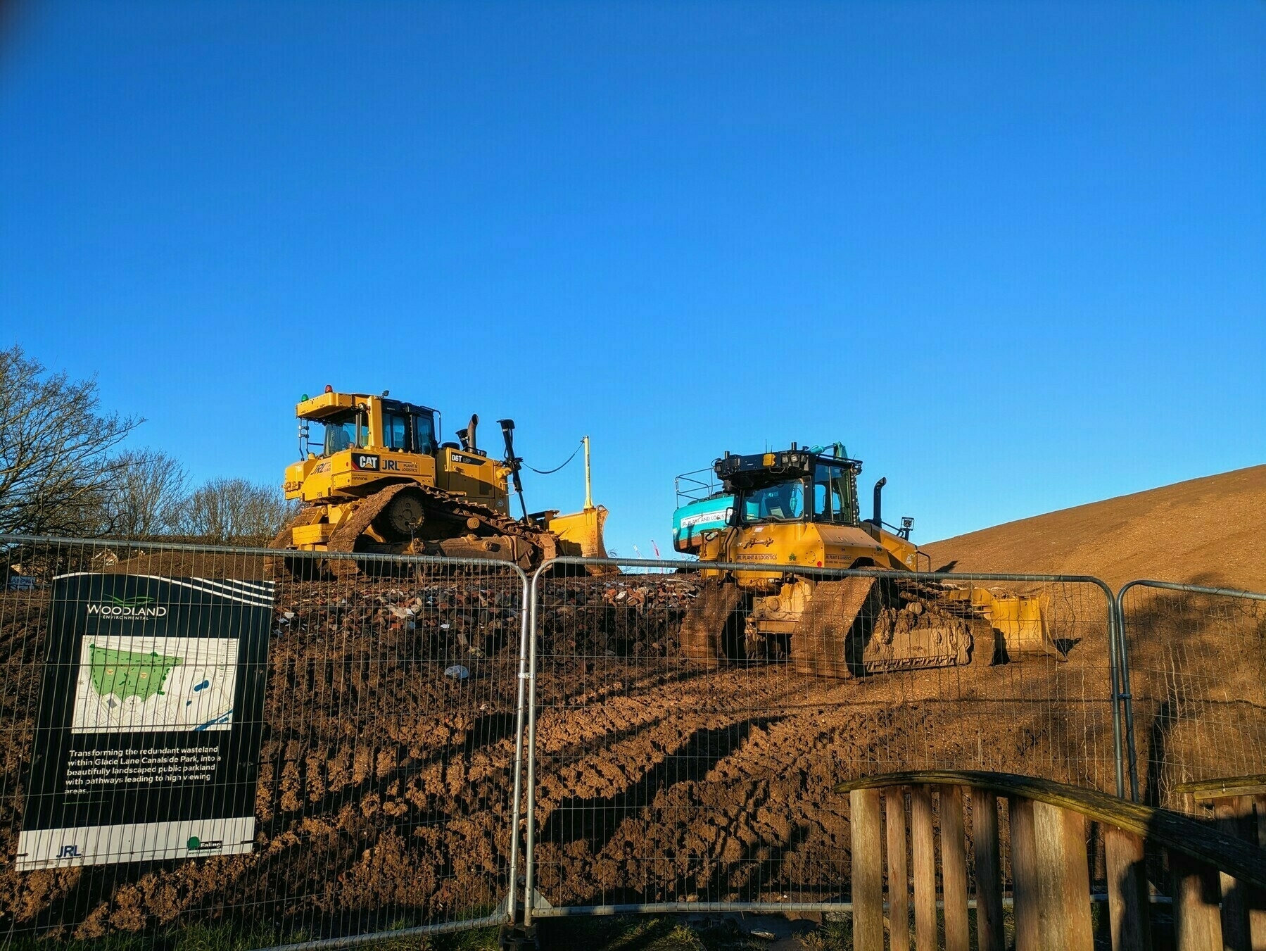 Two yellow bulldozers work on a dirt hill behind a metal fence under a clear blue sky.