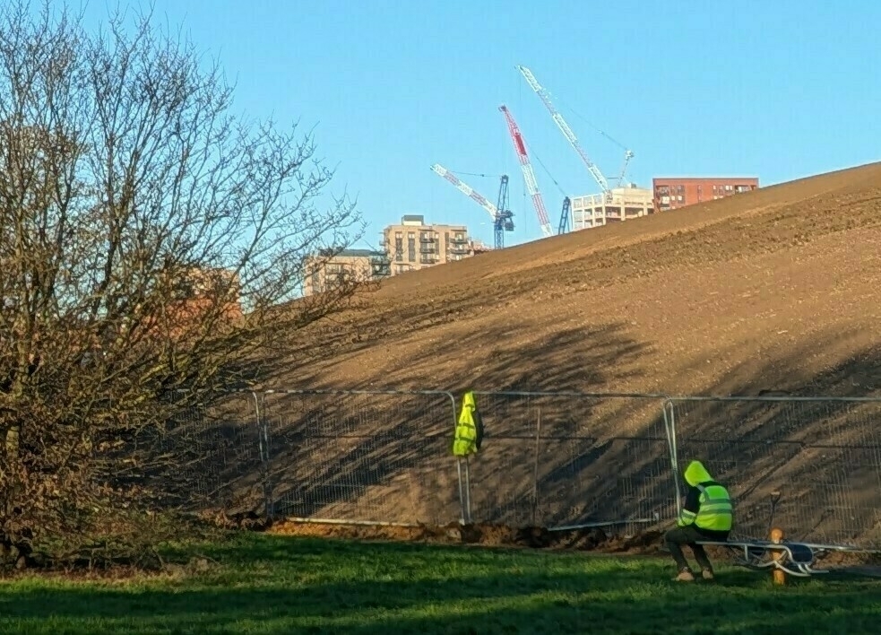 A person in a high-visibility jacket sits on a bench near a construction site with cranes in the background.