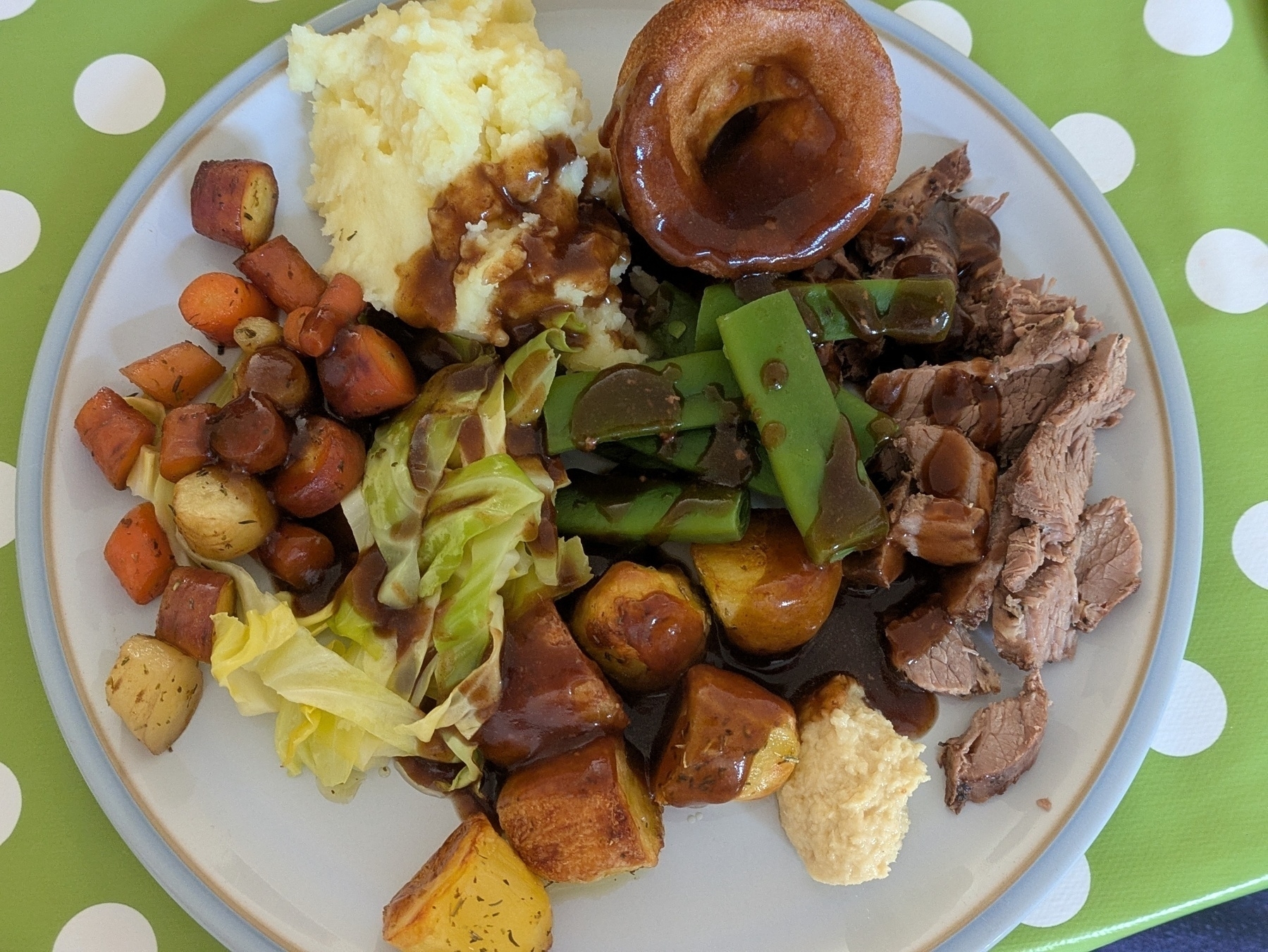 A plate of food featuring sliced roast meat, mashed potatoes, Yorkshire pudding, roasted vegetables, green beans, horseradish sauce and gravy.