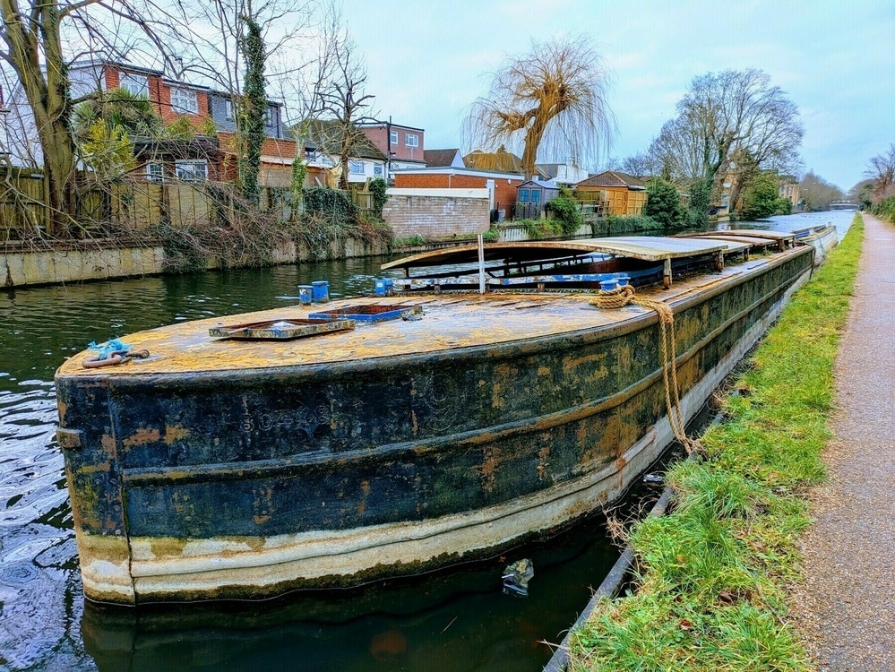 A large, weathered barge is moored alongside a canal path, surrounded by trees and residential buildings.