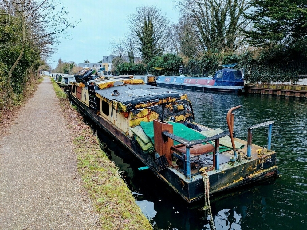 A weathered, dilapidated houseboat is moored along a narrow canal, with a path and trees lining the opposite bank.