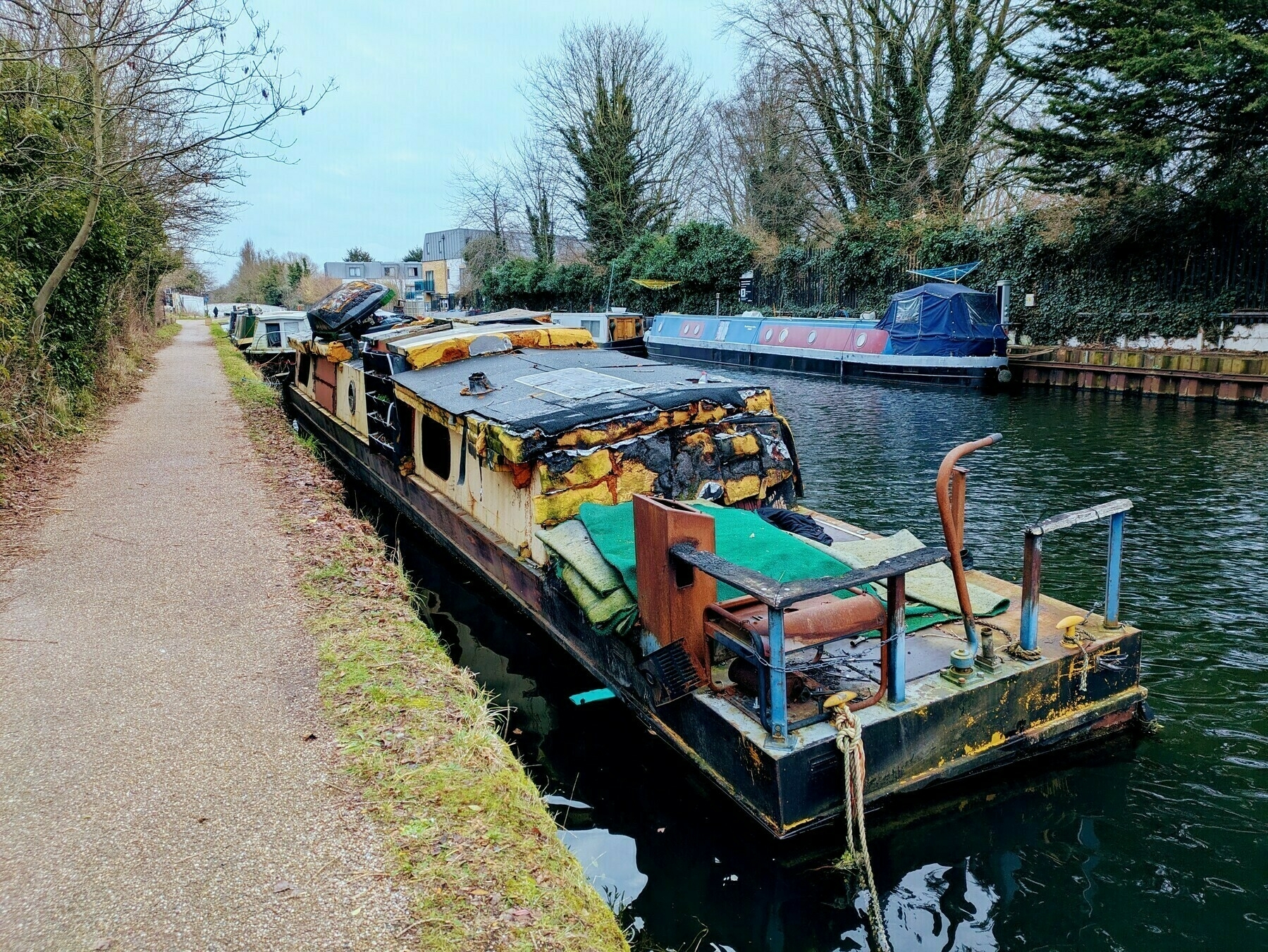 Auto-generated description: A weathered, dilapidated houseboat is moored along a narrow canal, with a path and trees lining the opposite bank.