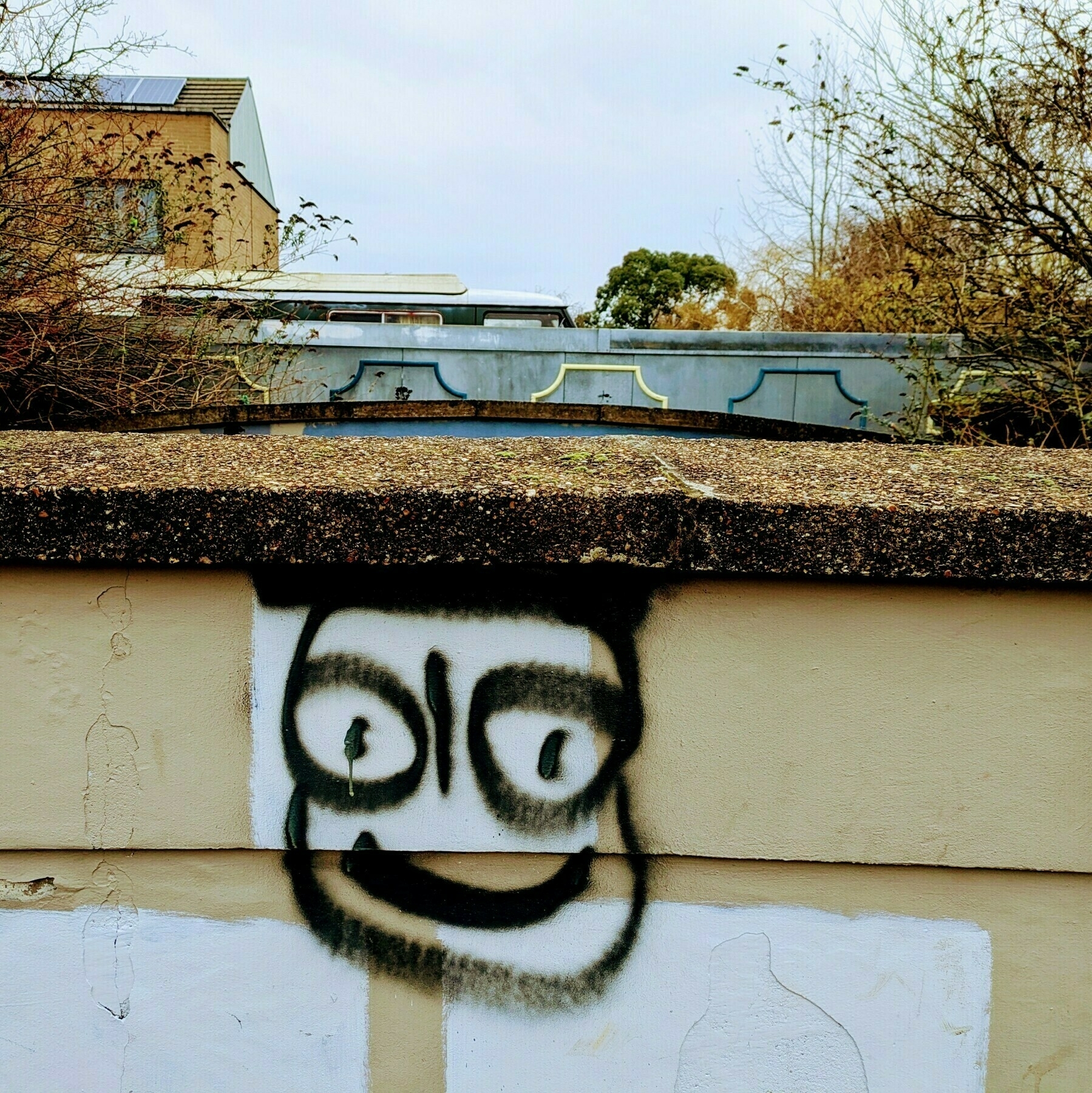 Auto-generated description: A wall facing the camera is painted with a simplistic, smiling face in black and white graffiti, set against an urban backdrop with trees and buildings.