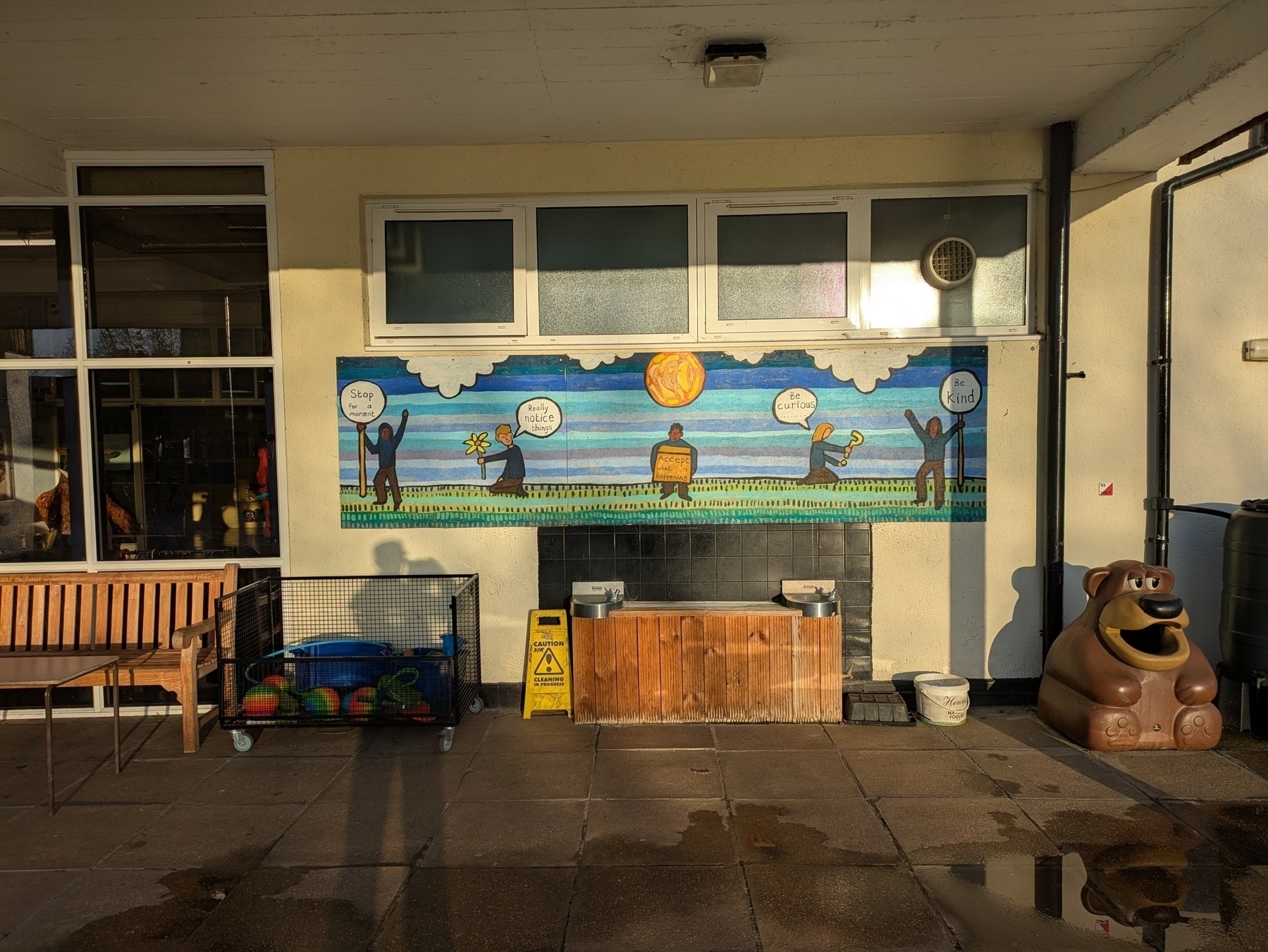 Auto-generated description: A colorful mural featuring cartoon figures is displayed on a school wall surrounded by various outdoor furnishings and play equipment.