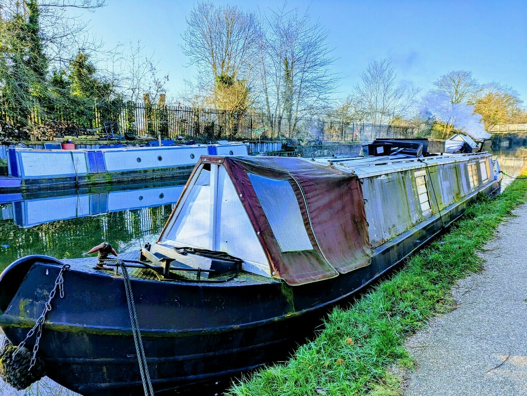 A narrowboat with a worn exterior is moored along a canal on a sunny day, smoke drifting upwards at the rear.