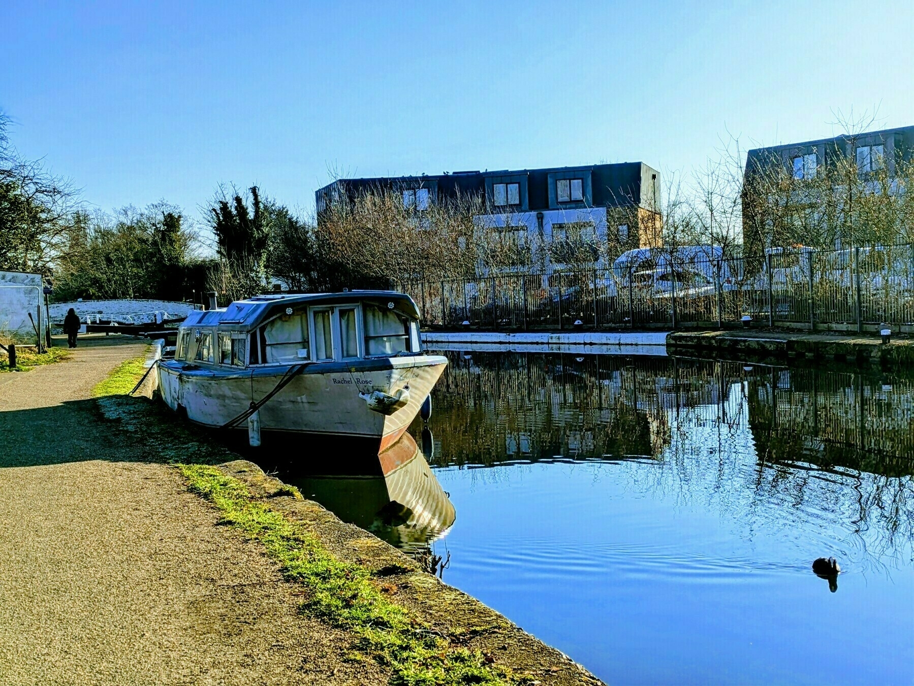 A boat is moored along a tranquil canal lined with trees and houses, under a clear blue sky.