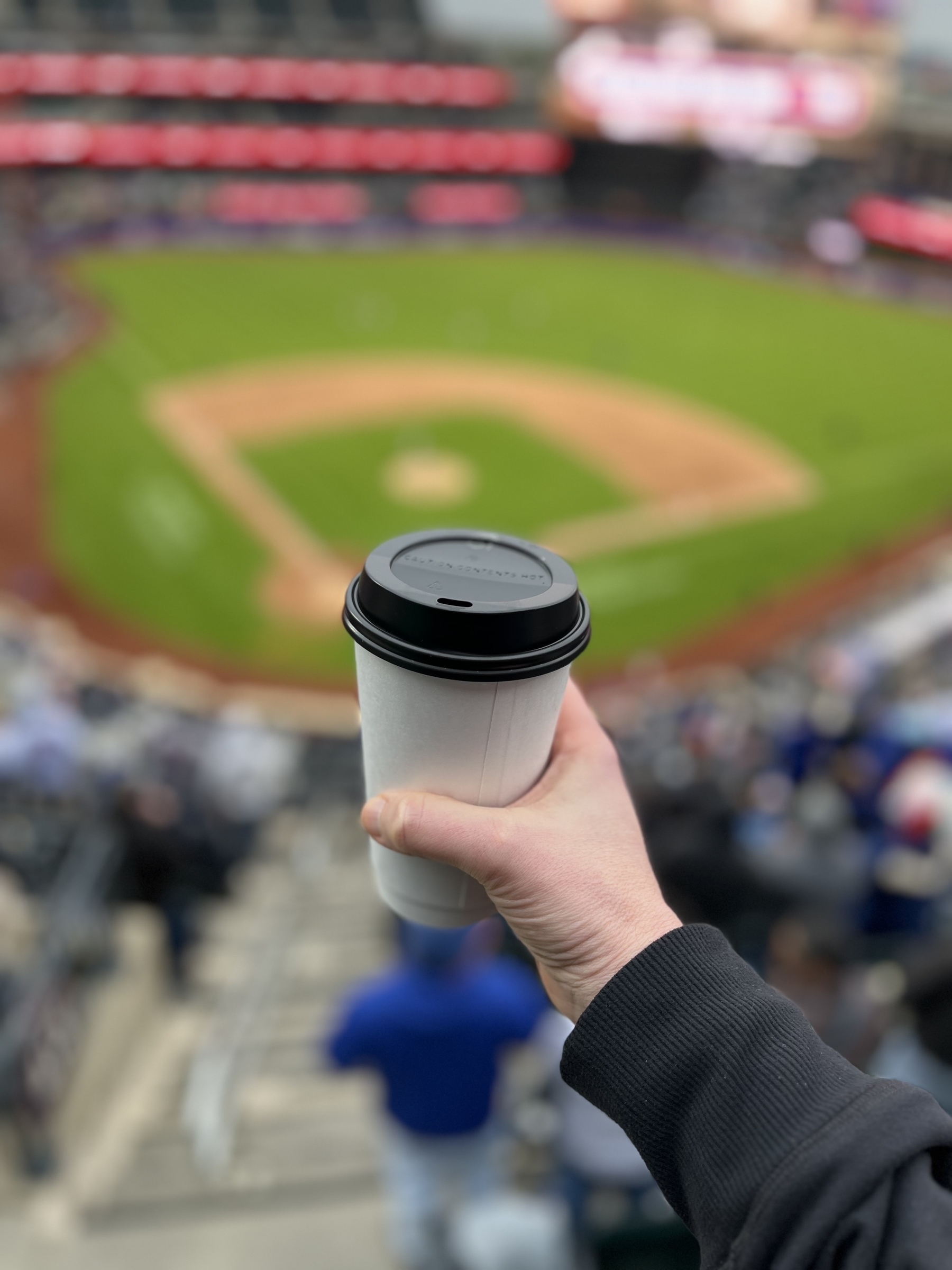 Auto-generated description: A person holds a coffee cup with a baseball field blurred in the background.