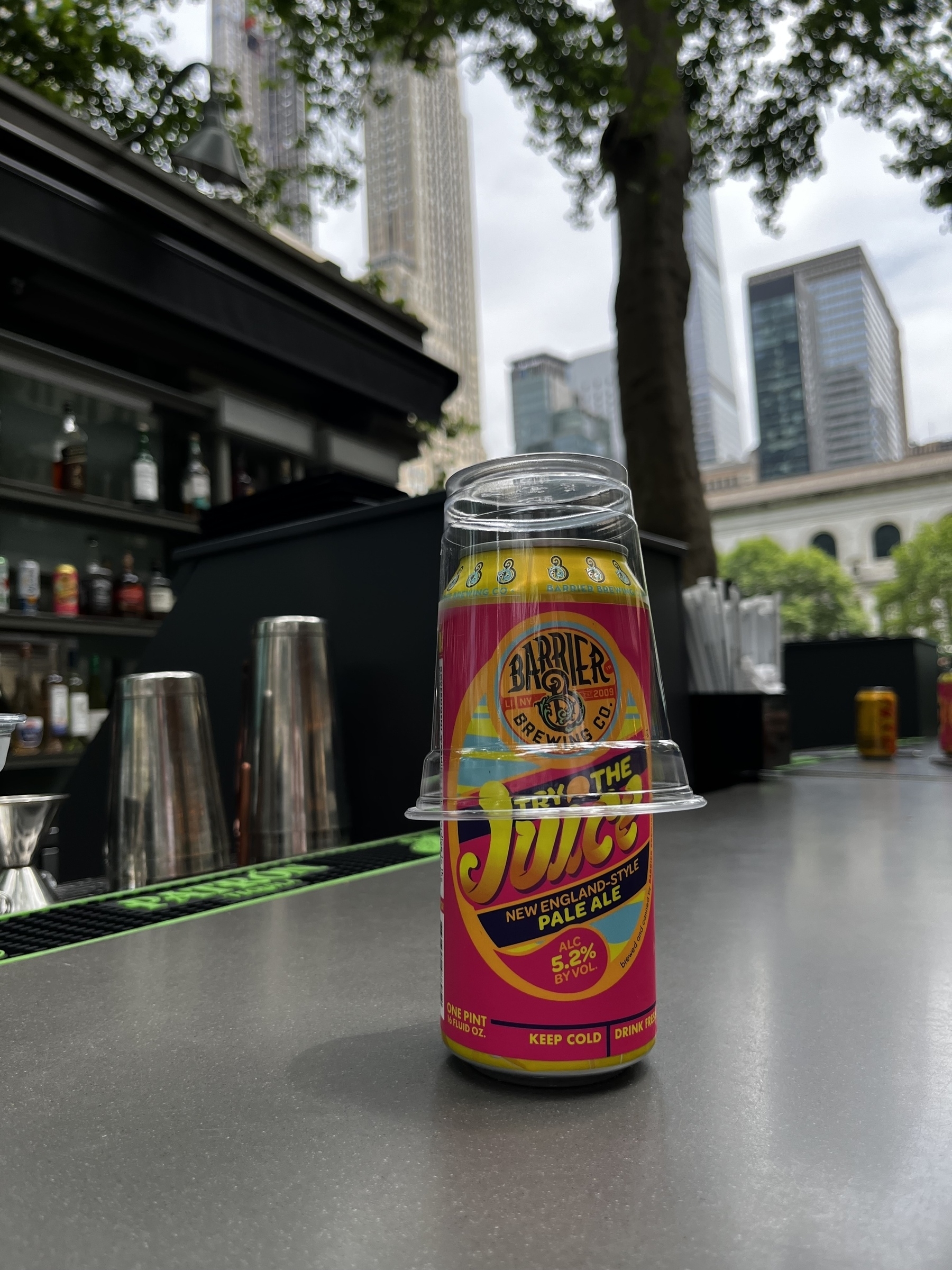 Auto-generated description: A can of Barrier Brewing Co’s Kickin' Knowledge Juice pale ale is placed on a bar counter, partially covered with a plastic cup, amidst an urban outdoor setting.
