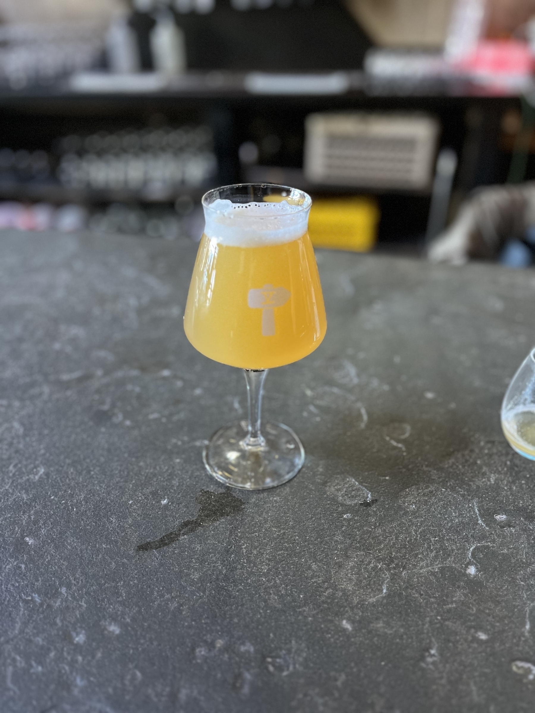 Auto-generated description: A tulip-shaped glass filled with a hazy, frothy beverage, possibly beer, sits on a dark countertop.
