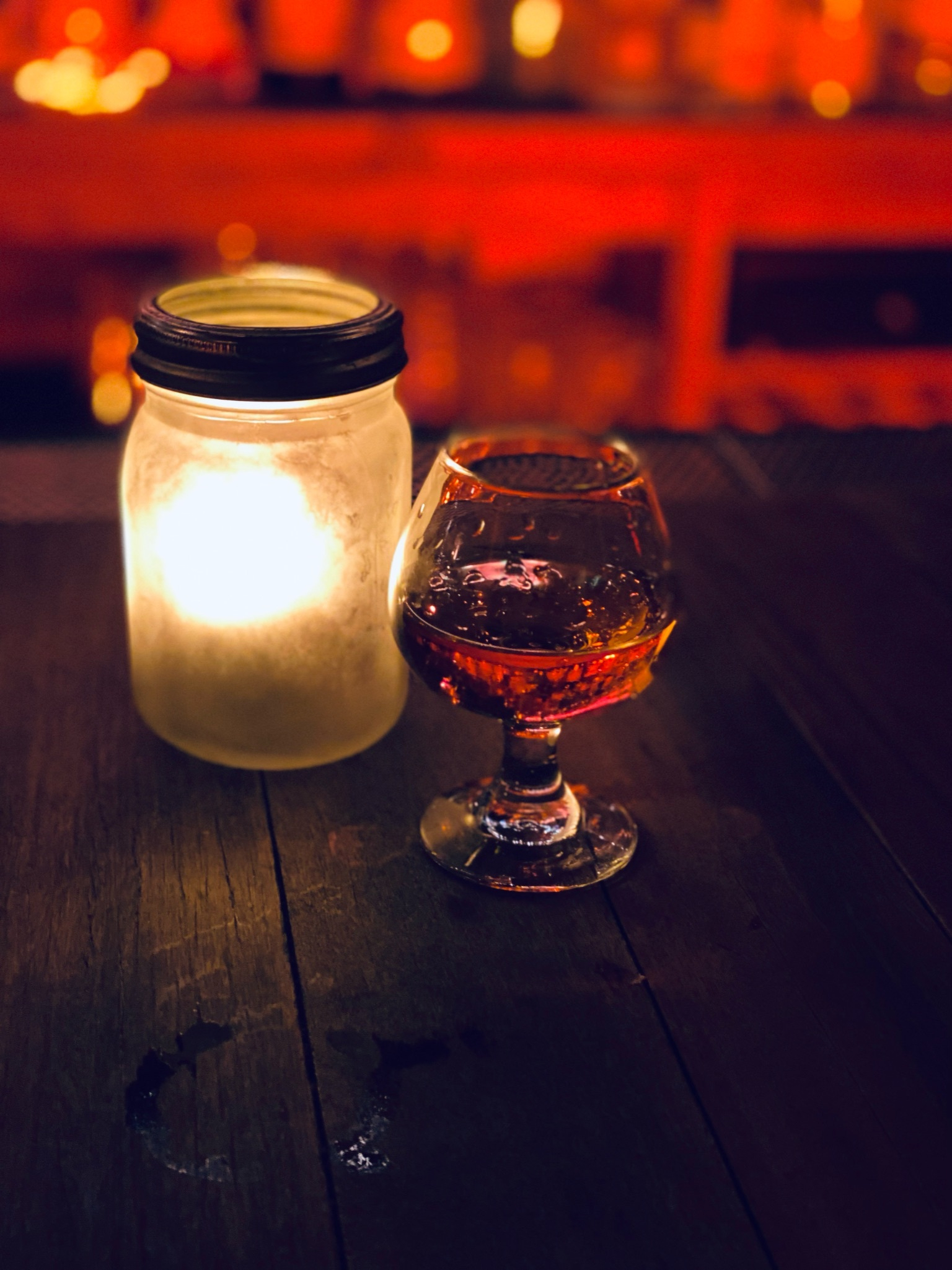 A lit candle in a frosted jar and a glass of amber liquid sit side by side on a wooden table, casting a warm glow.
