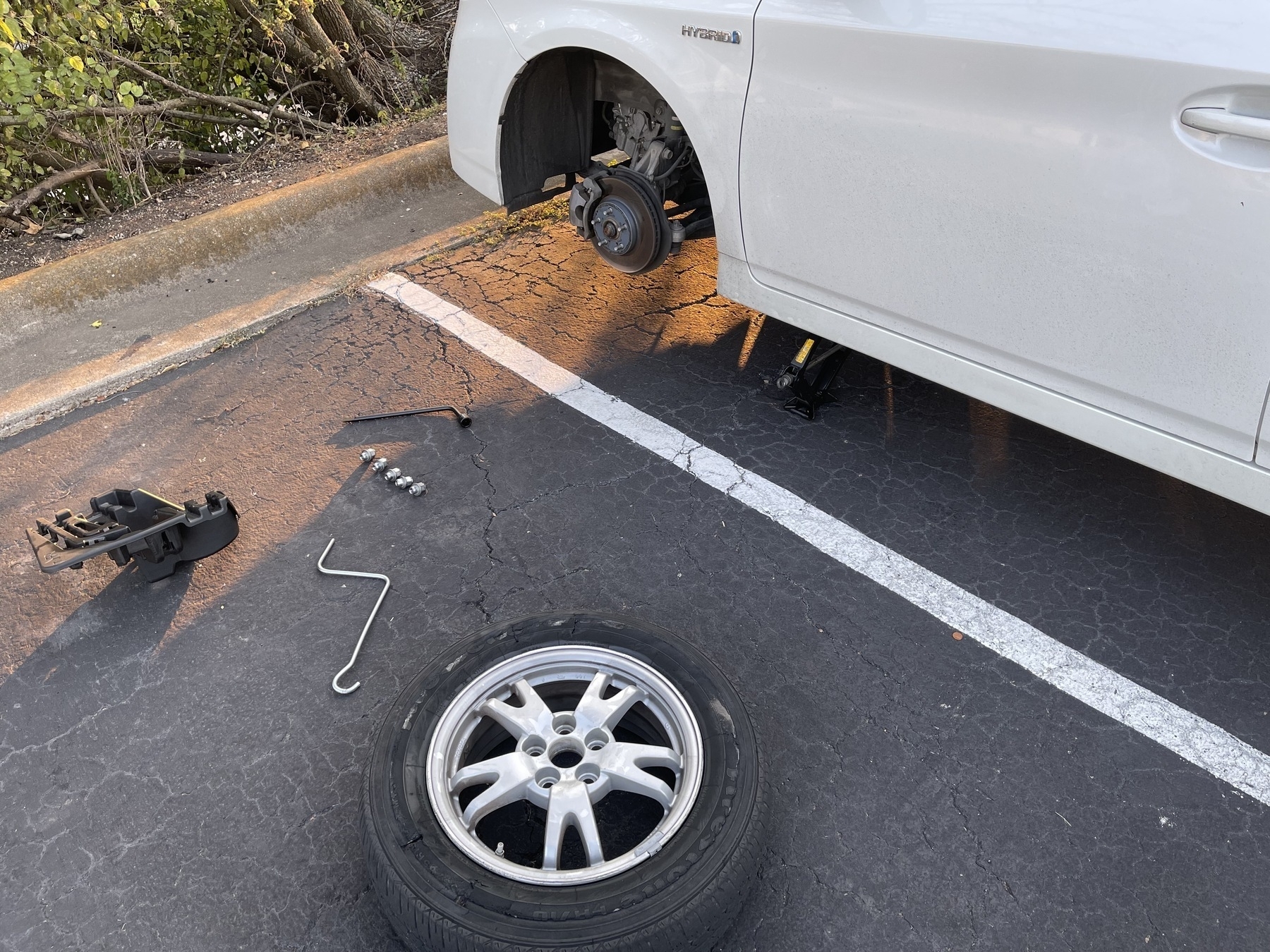 The front left side of a white Toyota Prius is shown on a jack with its tire removed. The tire is down on the next parking spot over, showing damage. Various tools are next to the tire on the ground.