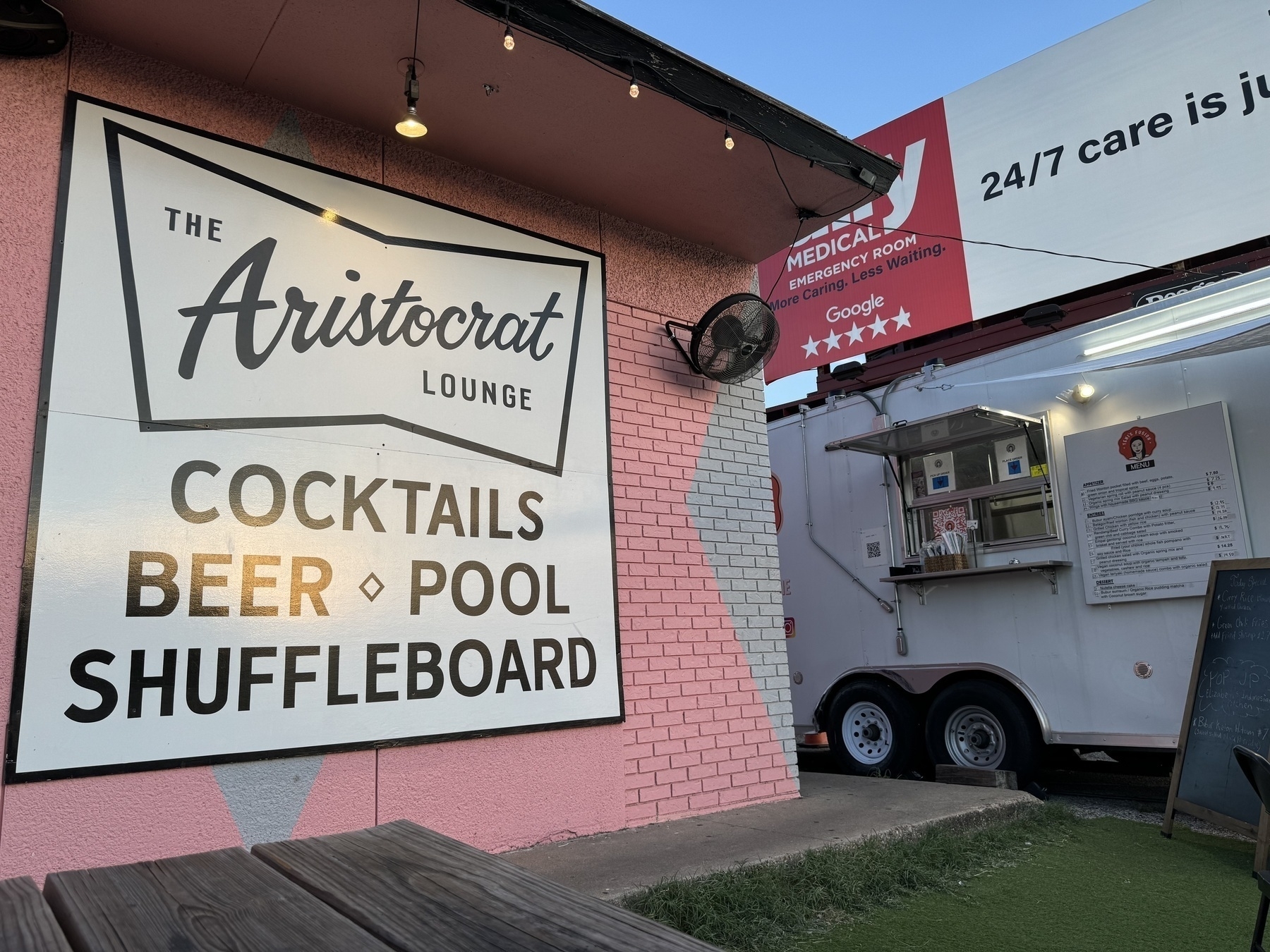 In the foreground, a sign for The Aristocrat Lounge / Cocktails, Beer, Pool, and Shuffleboard. In the background, a food truck, Yeni’s Fusion.