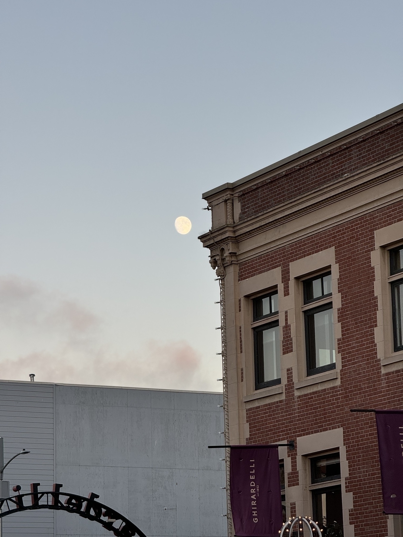 Photo shows a blue sky with an almost full moon and the old Ghirardelli building 