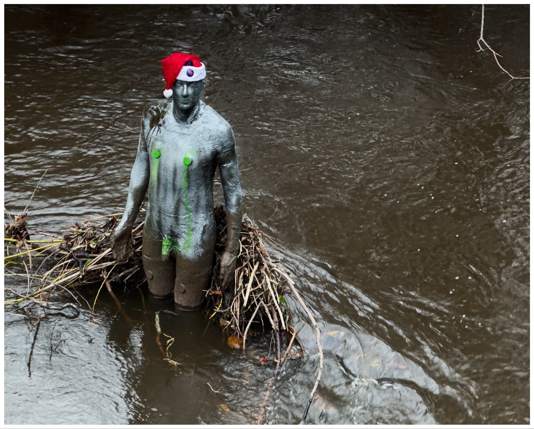 Statue of a person wearing a Santa hat, partially submerged in a river, with green algae-like markings on the body.