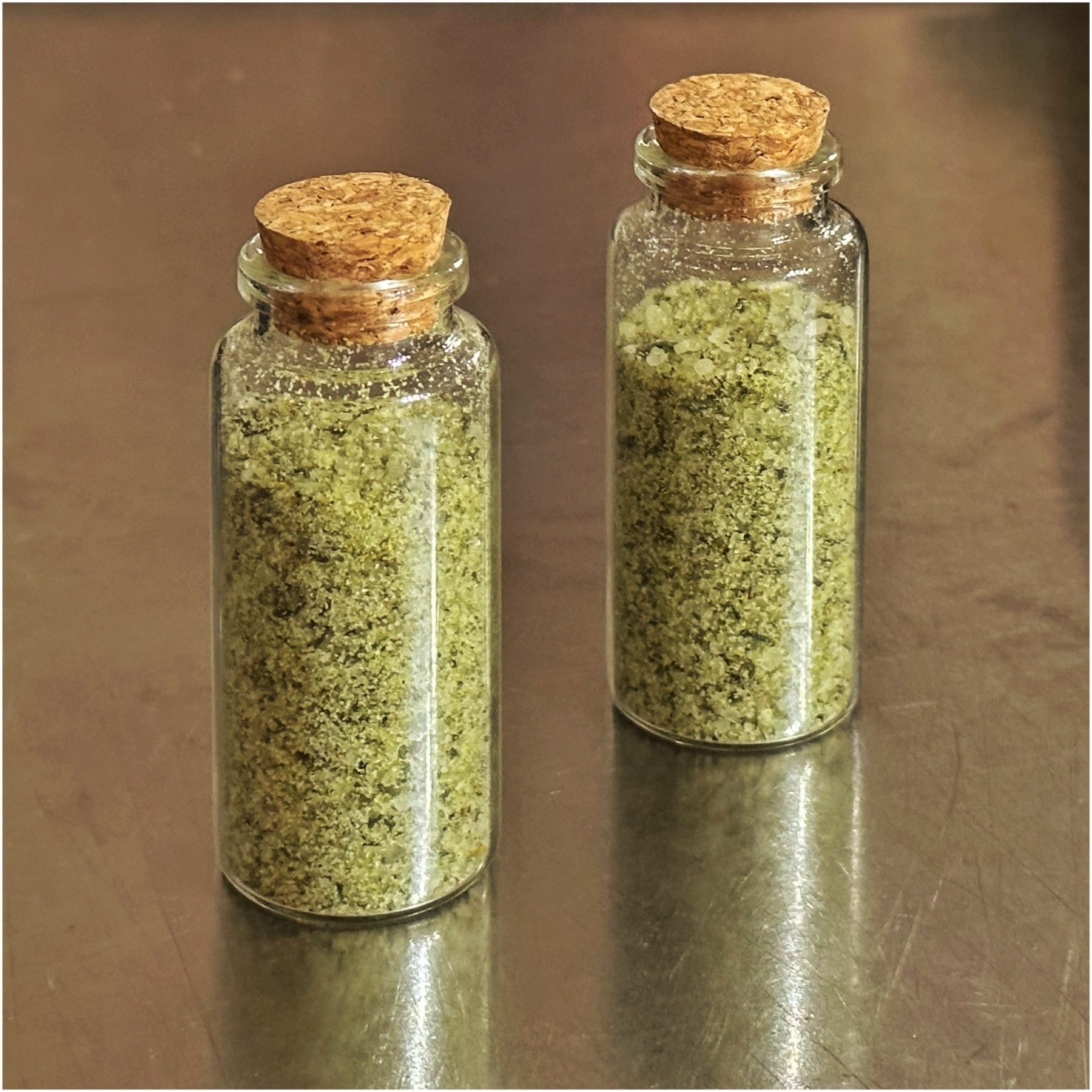 Two glass bottles with cork stoppers filled with a green herbal substance, possibly a spice or seasoning, standing on a metallic surface.