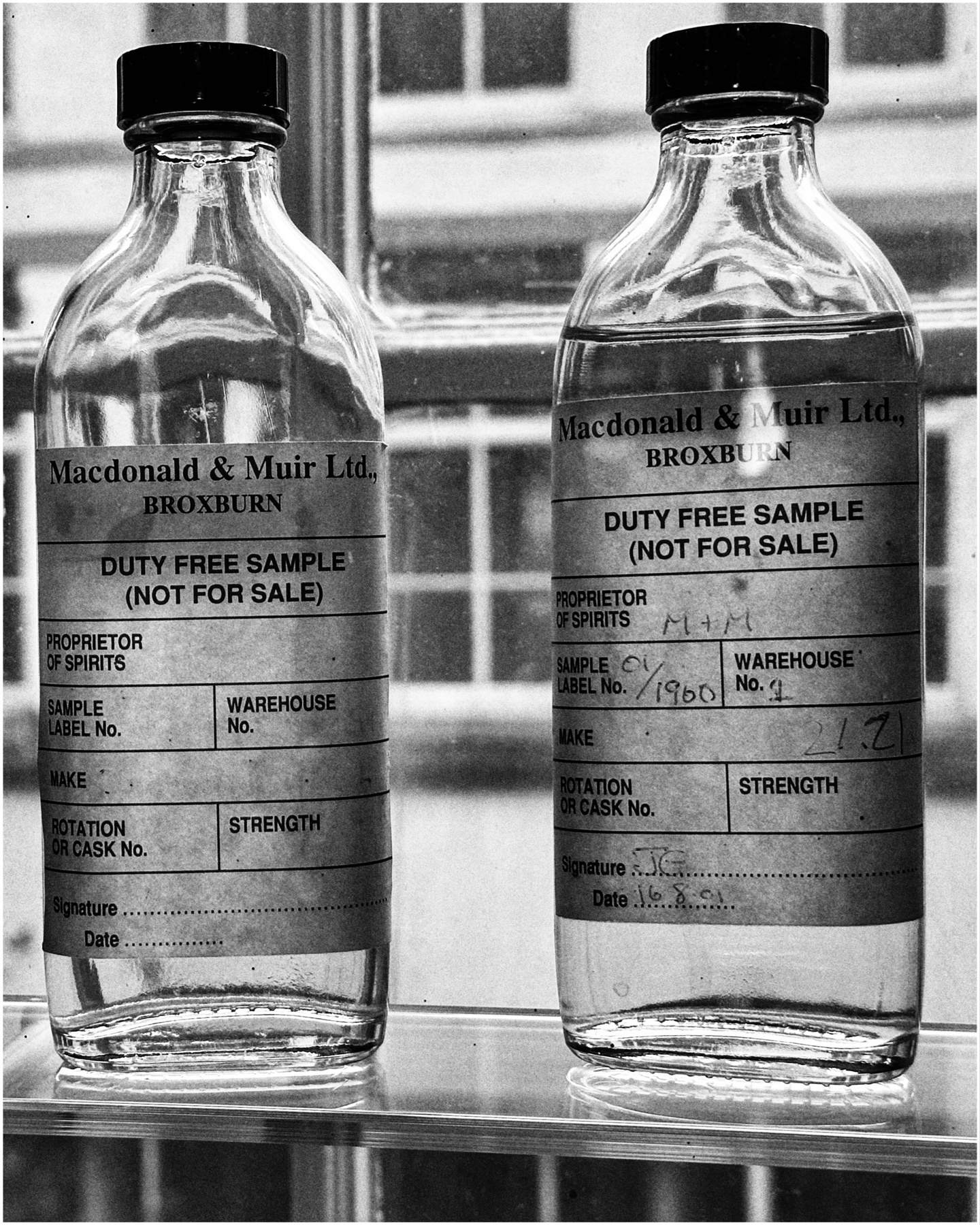 Two glass bottles with black caps, labeled "Macdonald & Muir Ltd, Broxburn," marked as "DUTY FREE SAMPLE (NOT FOR SALE)," with additional details filled out in handwriting. They are set against a window