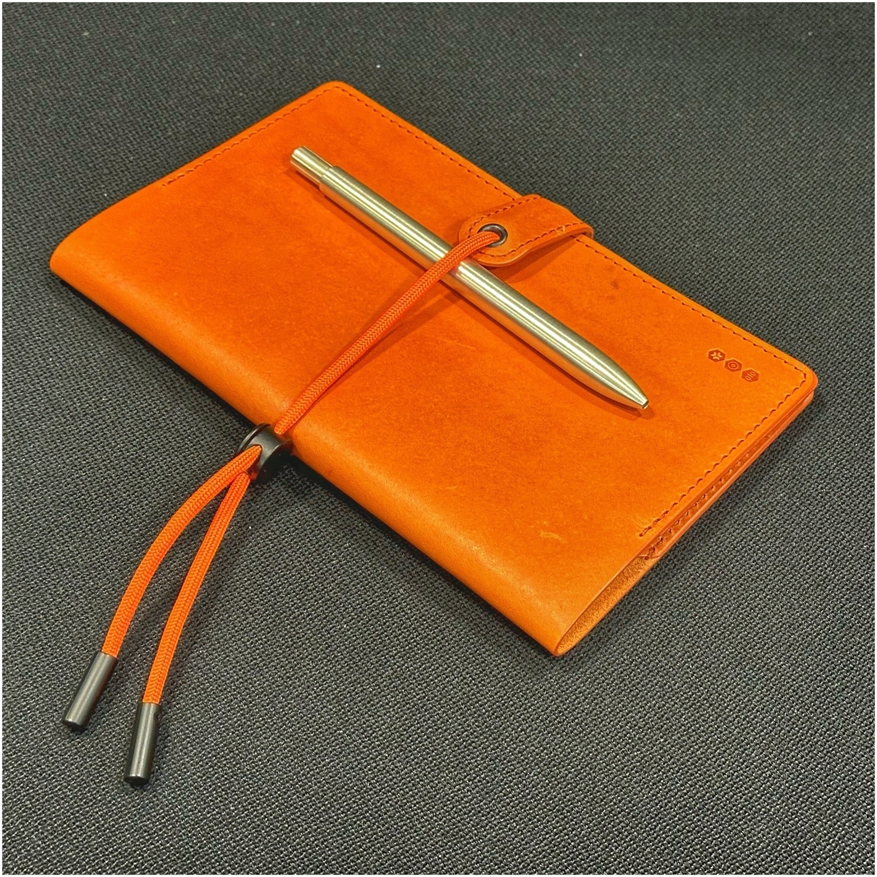 Orange journal with an elastic closure and a silver pen resting on top, placed on a dark textured surface.