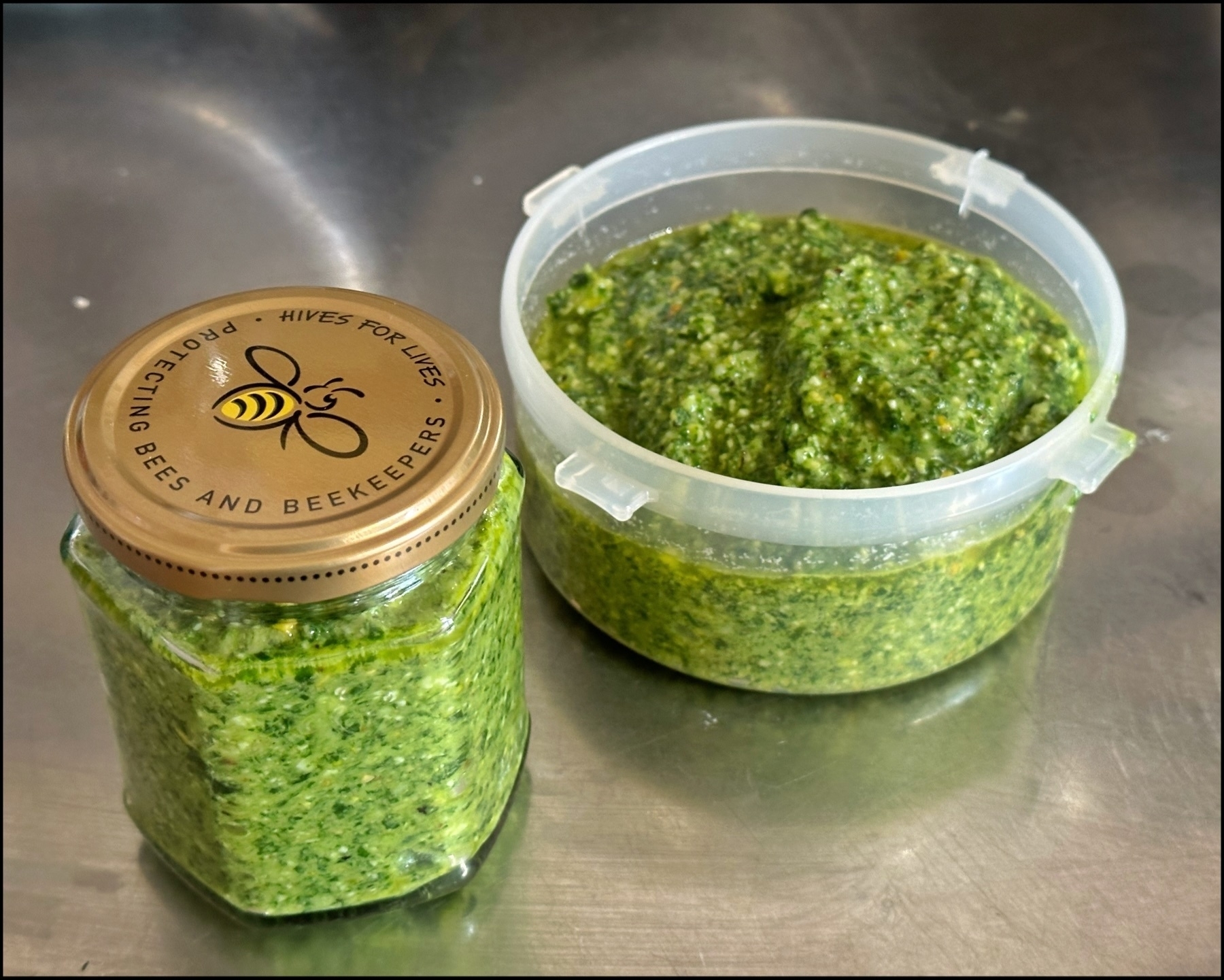 Jar of pesto with a bee-themed lid alongside an open plastic container of pesto on a stainless steel surface.