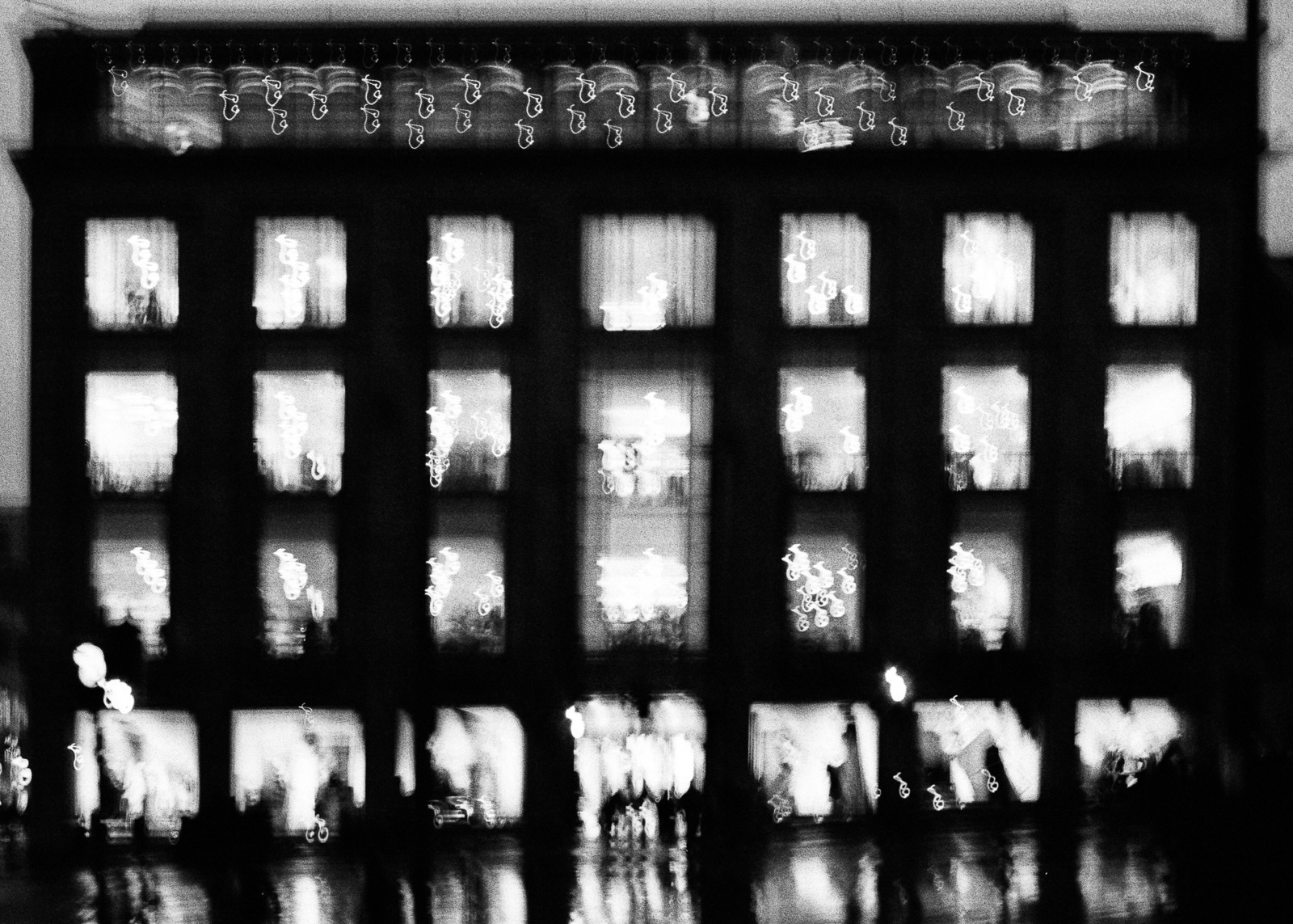 Black and white image of a blurred building facade at night with multiple windows showing interior lights, giving a bokeh effect.