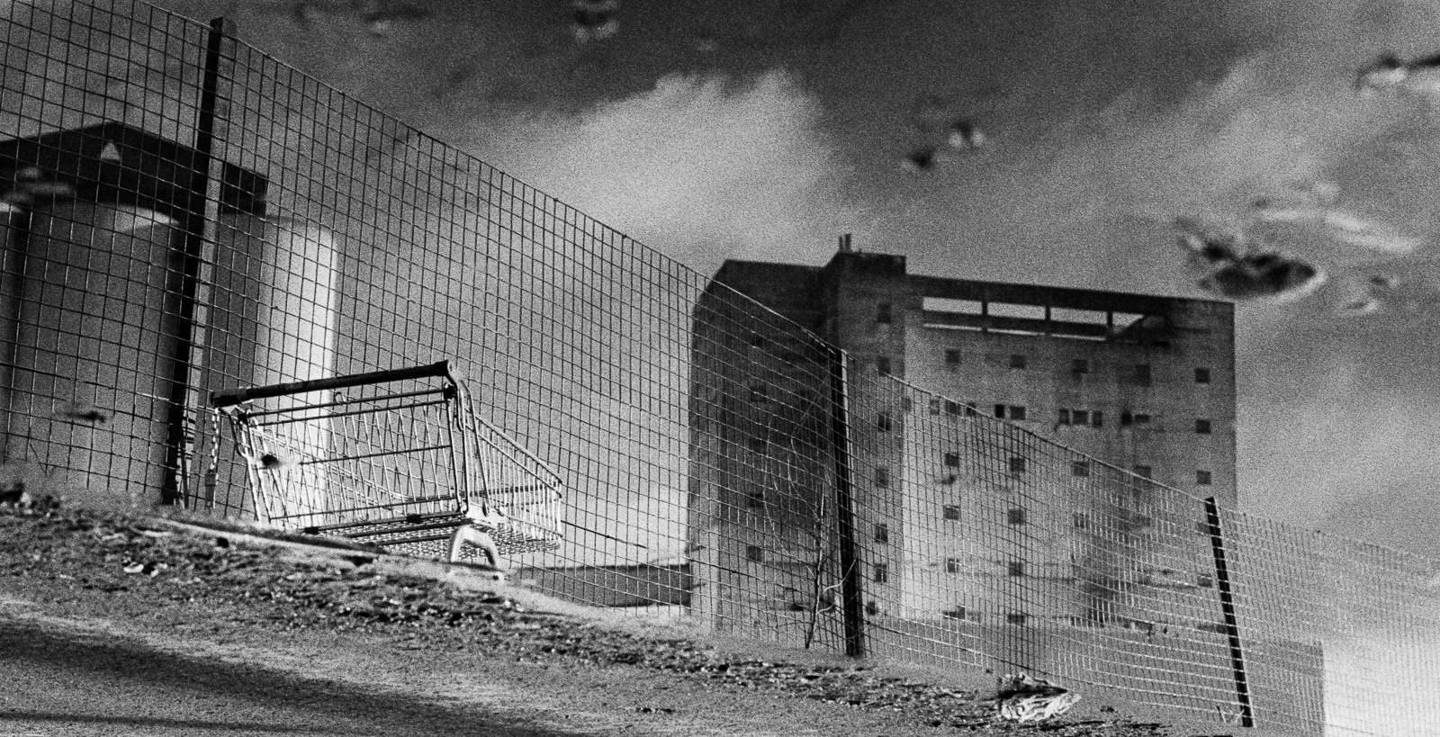 A black and white photograph showing a distorted reflection of a building and a fence in a puddle, with a shopping cart lying on its side partially submerged in water. The sky and clouds appear in the background, inverted due to the reflection.