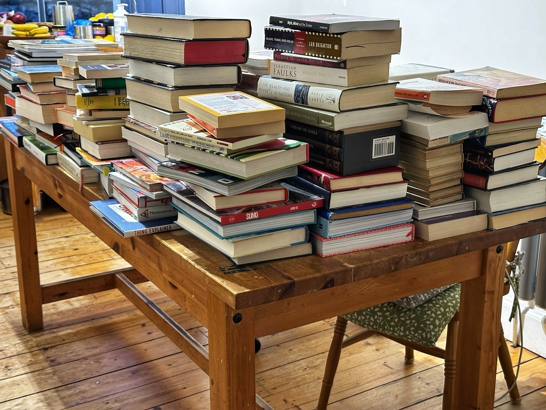 A large assortment of books stacked in multiple piles on a wooden table, with a wooden floor in the background.