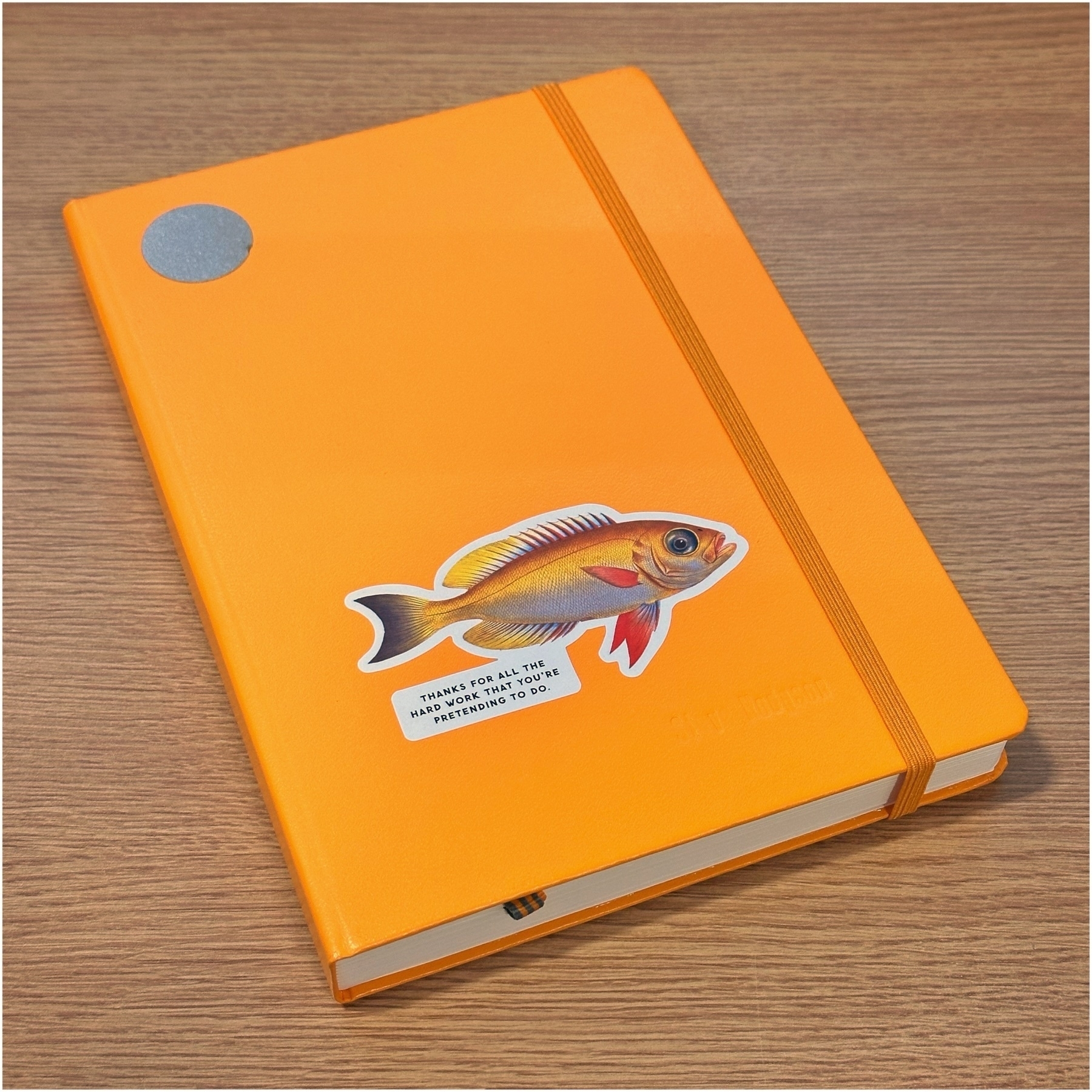 Orange notebook with an elastic closure and a fish illustration, featuring a humorous caption: "Thanks for all the hard work you're pretending to do."