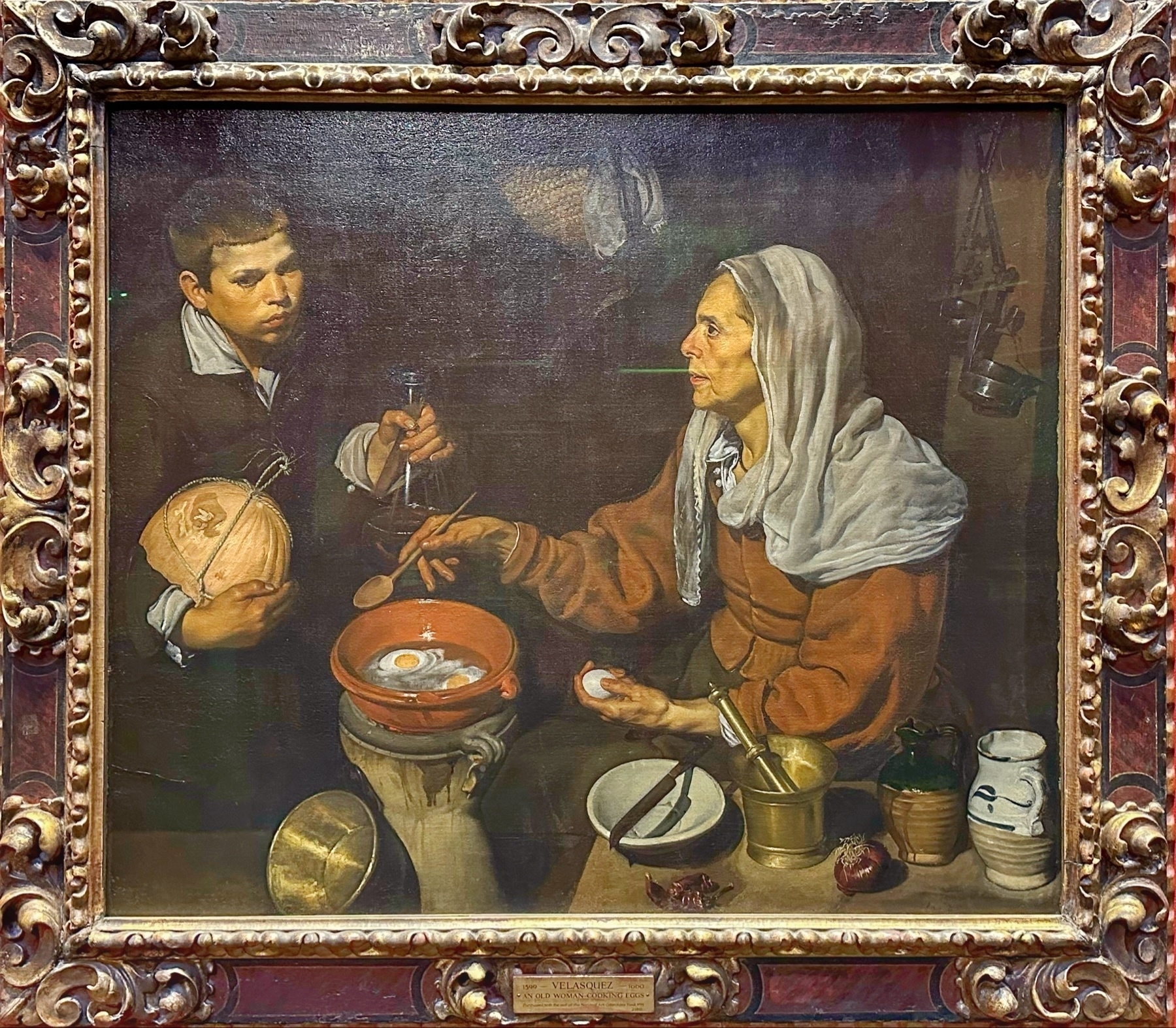 Oil painting depicting an older woman cooking eggs in a pan over a flame, with a young boy watching intently. They are surrounded by kitchen items including a mortar and pestle, a jug, and some vegetables.