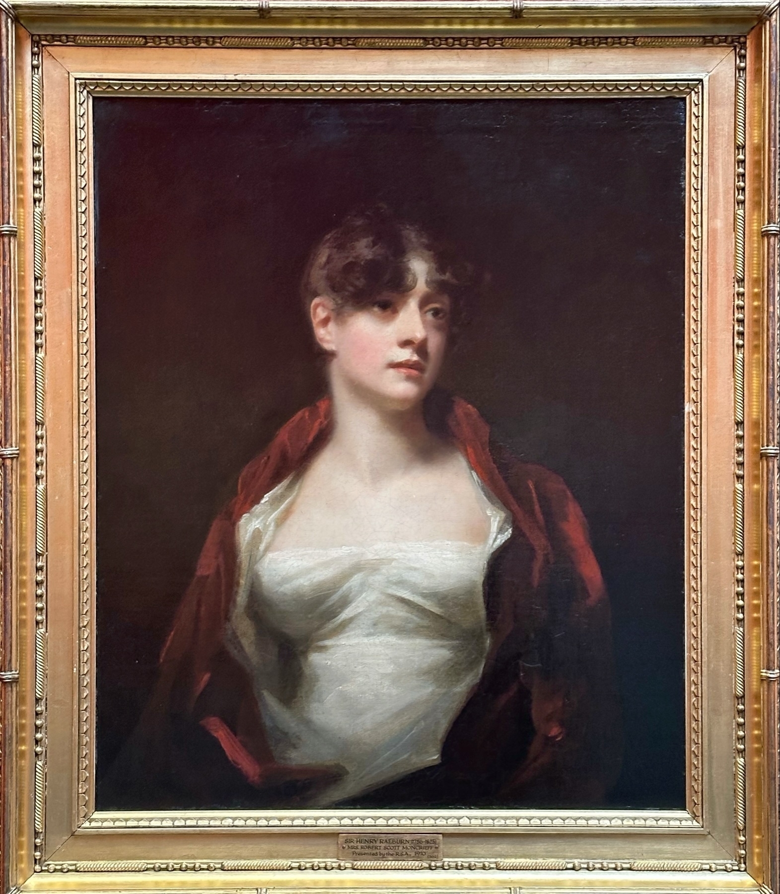 Oil painting of a young woman with dark hair wearing a white dress and a red draped shawl, set in an ornate gold frame. The woman is gazing off to the side with a soft expression.