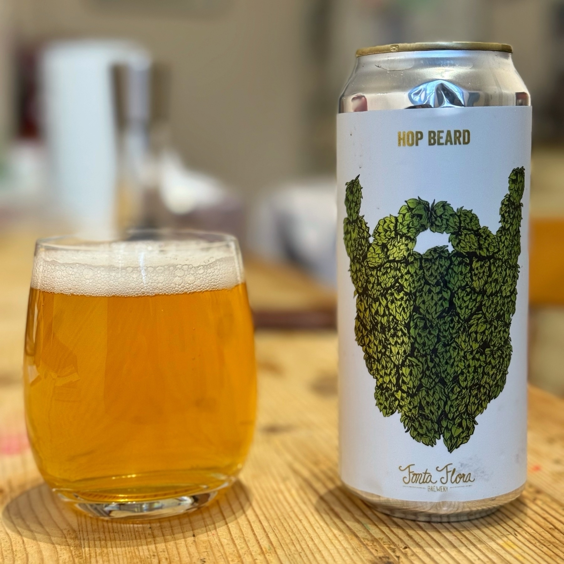 A can of "Hop Beard" beer from Fonta Flora Brewery is next to a glass filled with the beer. The can features an illustration of a beard made of hops. The background is slightly blurred.