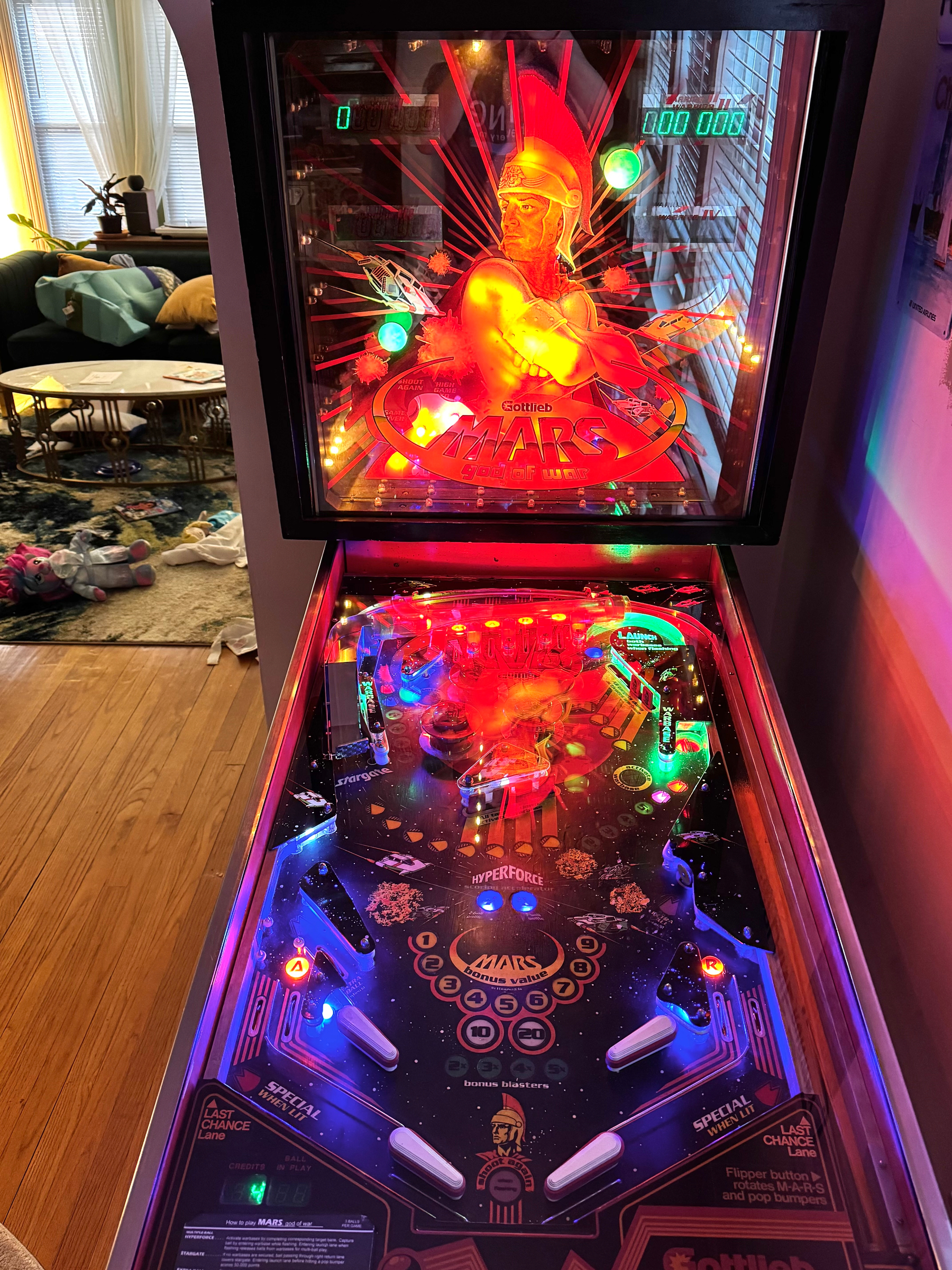 A colorful pinball machine with vibrant lights and graphics is set up in a cozy room with a visible couch and scattered items on the floor.