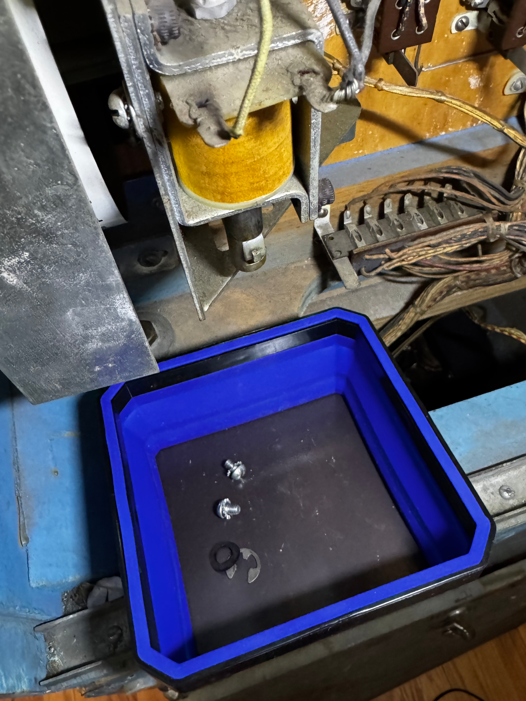 A blue tray containing small screws and a washer is positioned beneath a mechanical or electronic device with various wires and components.