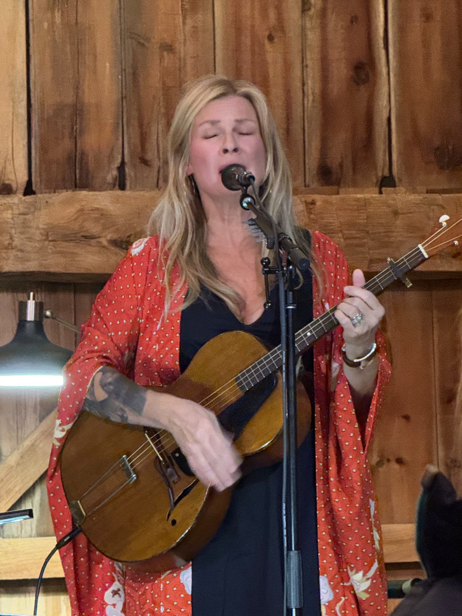 Karin Bergquist playing an acoustic guitar and singing into a microphone in the barn at Nowhere Else Farm