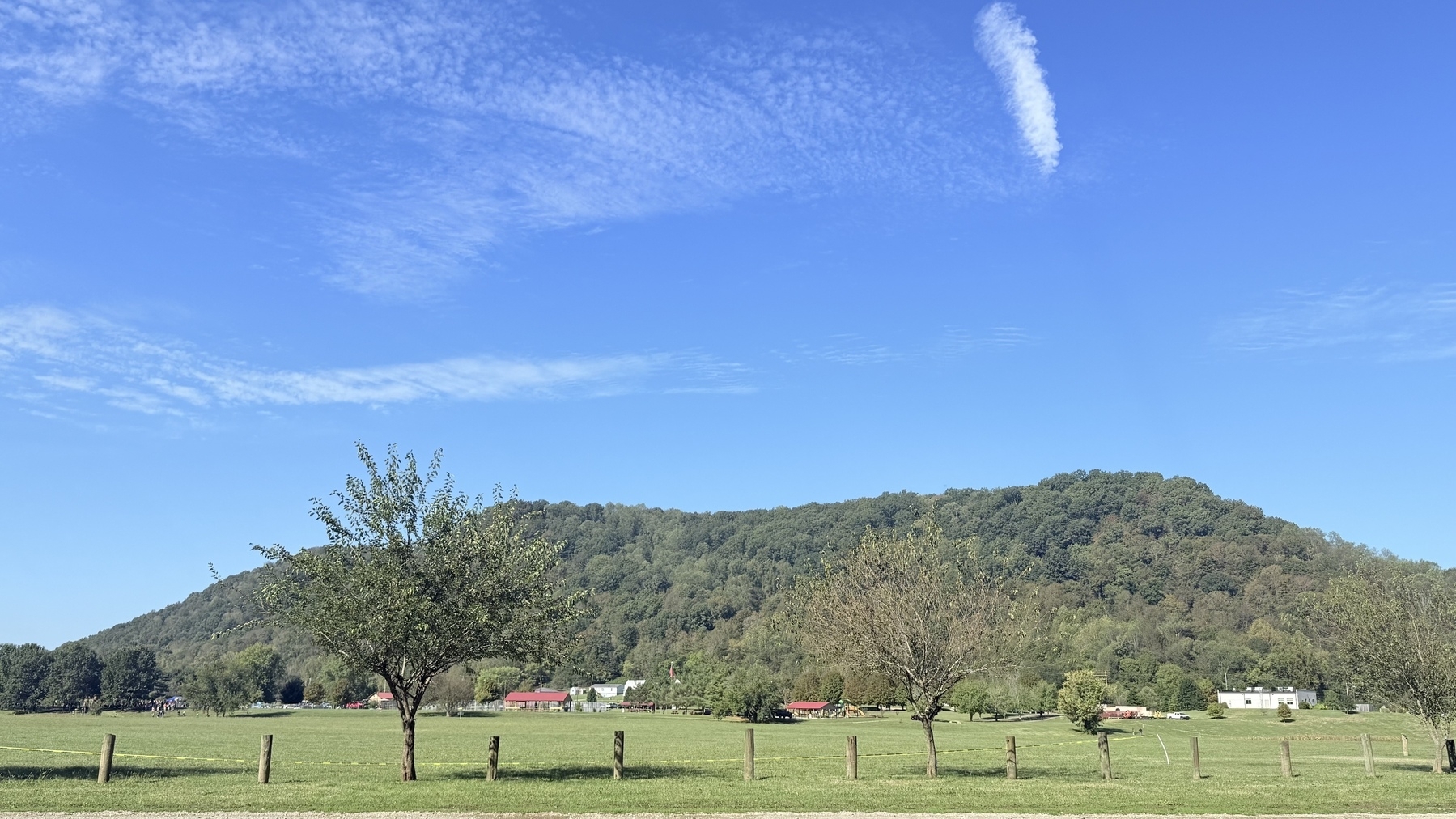 A picturesque landscape features a grassy field with trees, a distant hill, scattered houses, and a bright blue sky with wispy clouds.