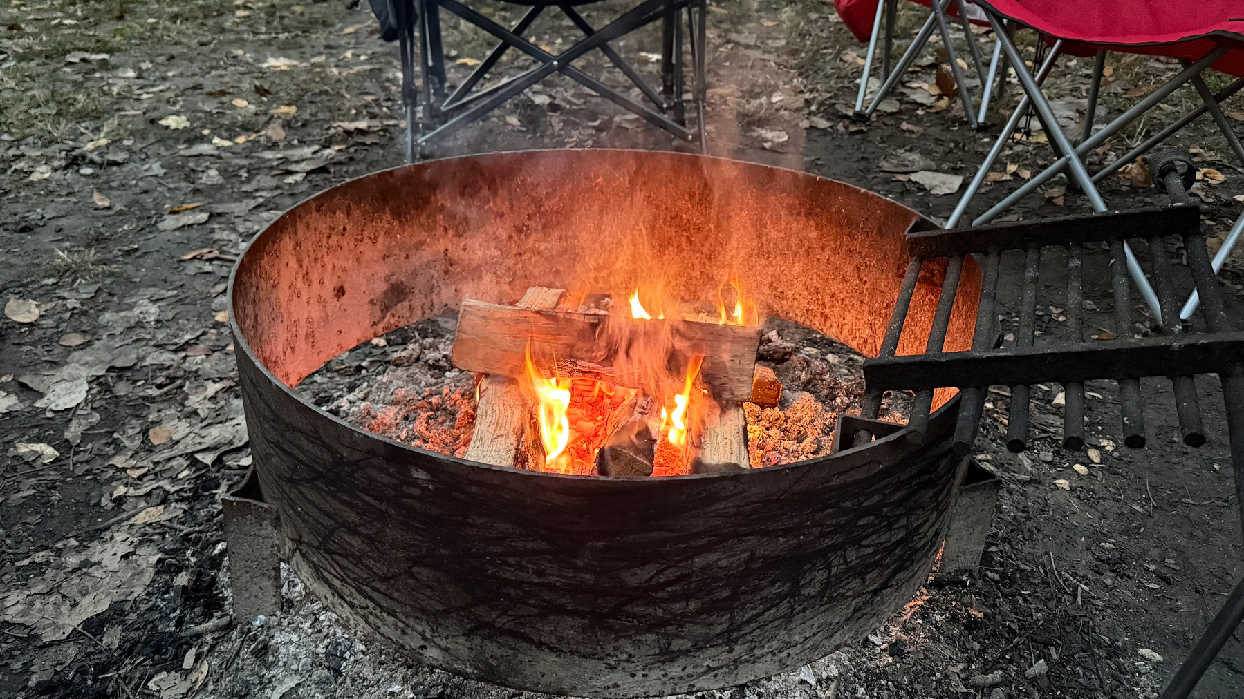 A campfire burns brightly in a metal fire pit surrounded by chairs on a dirt surface.&10;