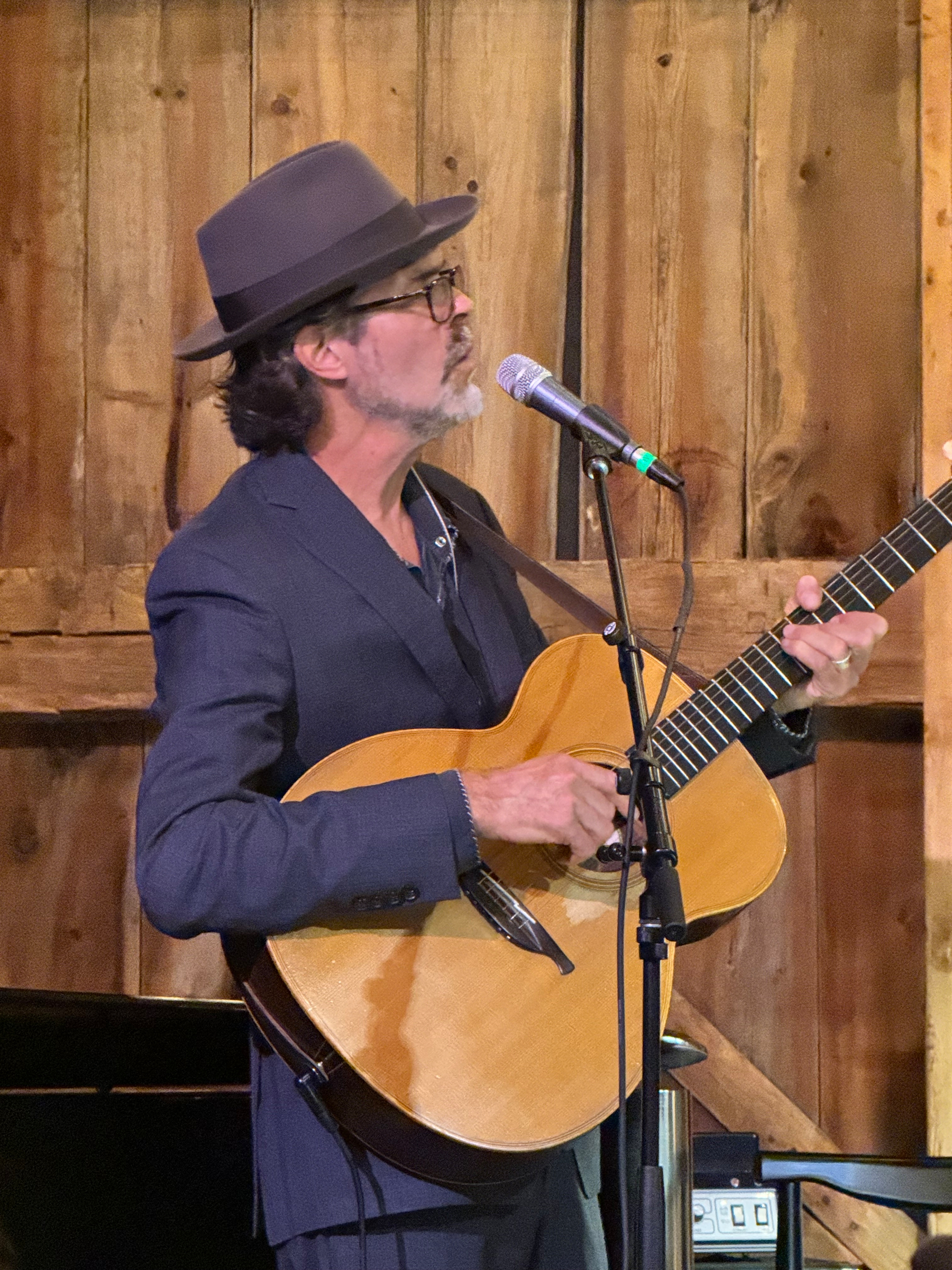Linford Detweiler playing an acoustic guitar while singing into a microphone in the barn at Nowhere Else Farm