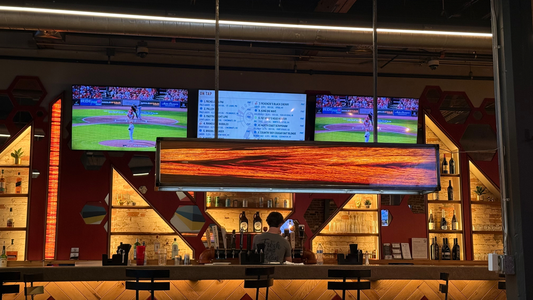 A bar with illuminated shelves, a bartender, and three screens displaying a Reds baseball game.