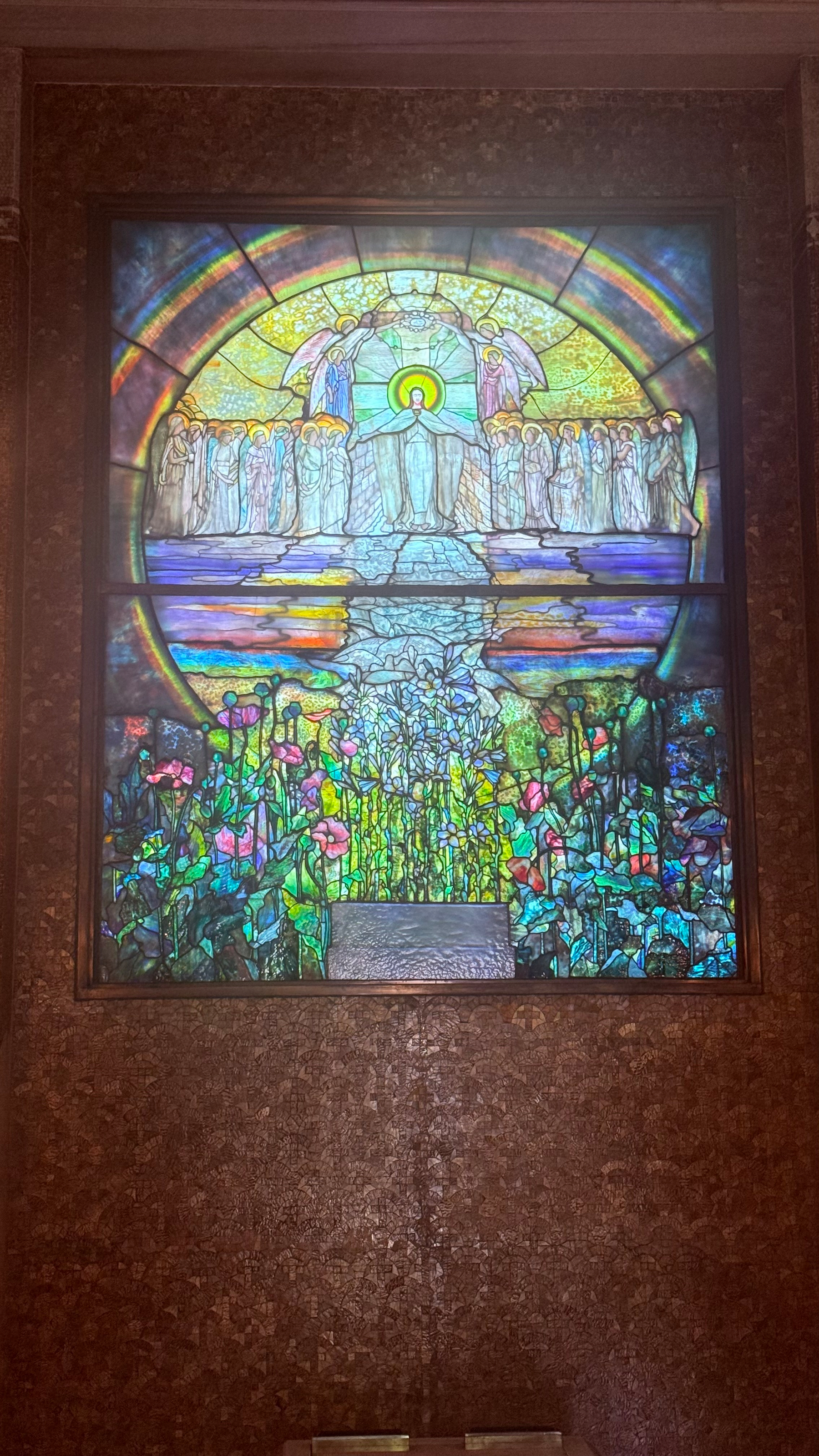 A vibrant stained glass window depicts a central angelic figure surrounded by other angels above a colorful garden filled with flowers.