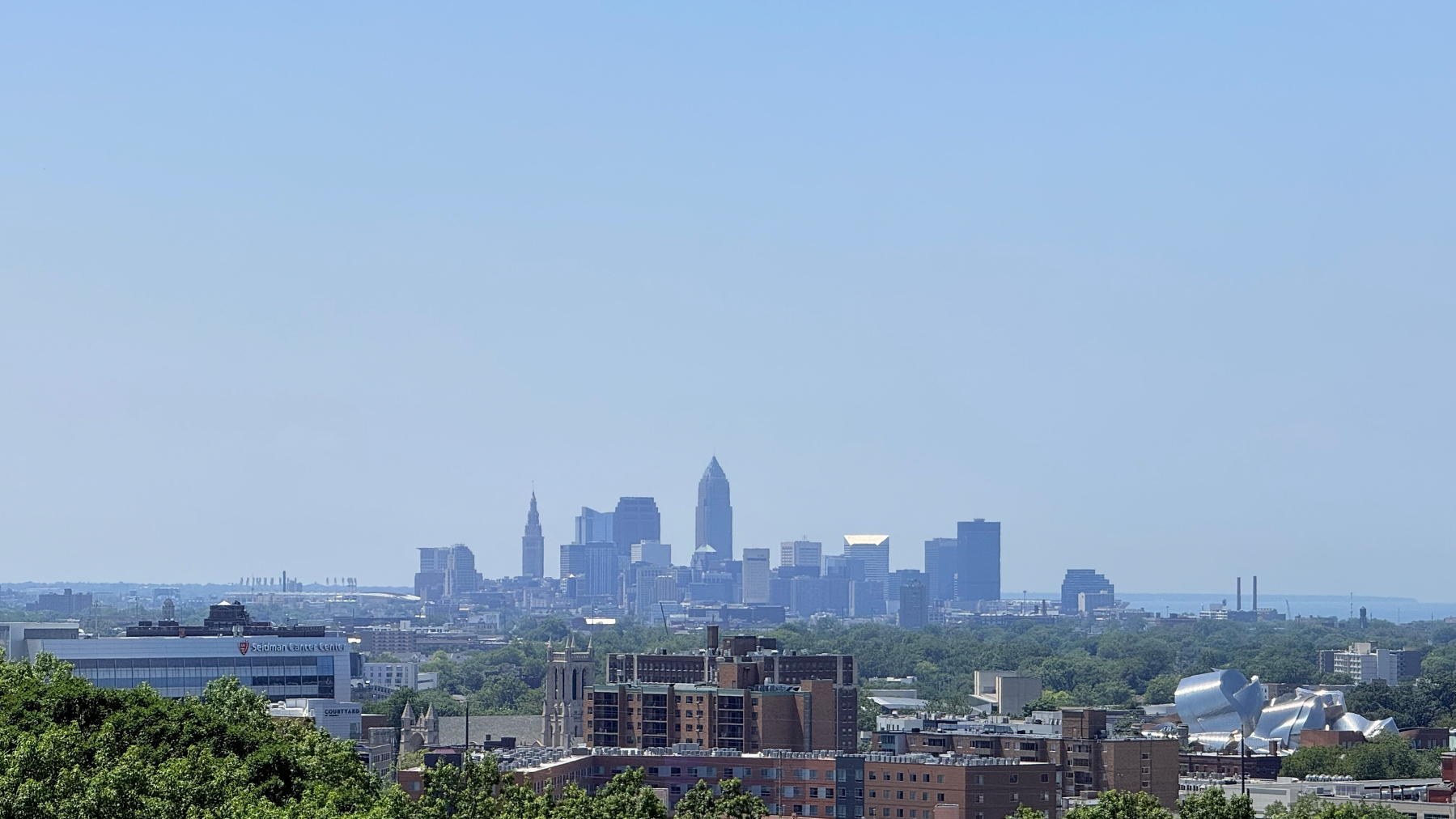The clear Cleveland  city skyline with a cluster of tall buildings rises above a foreground of shorter buildings and abundant greenery under a bright blue sky.