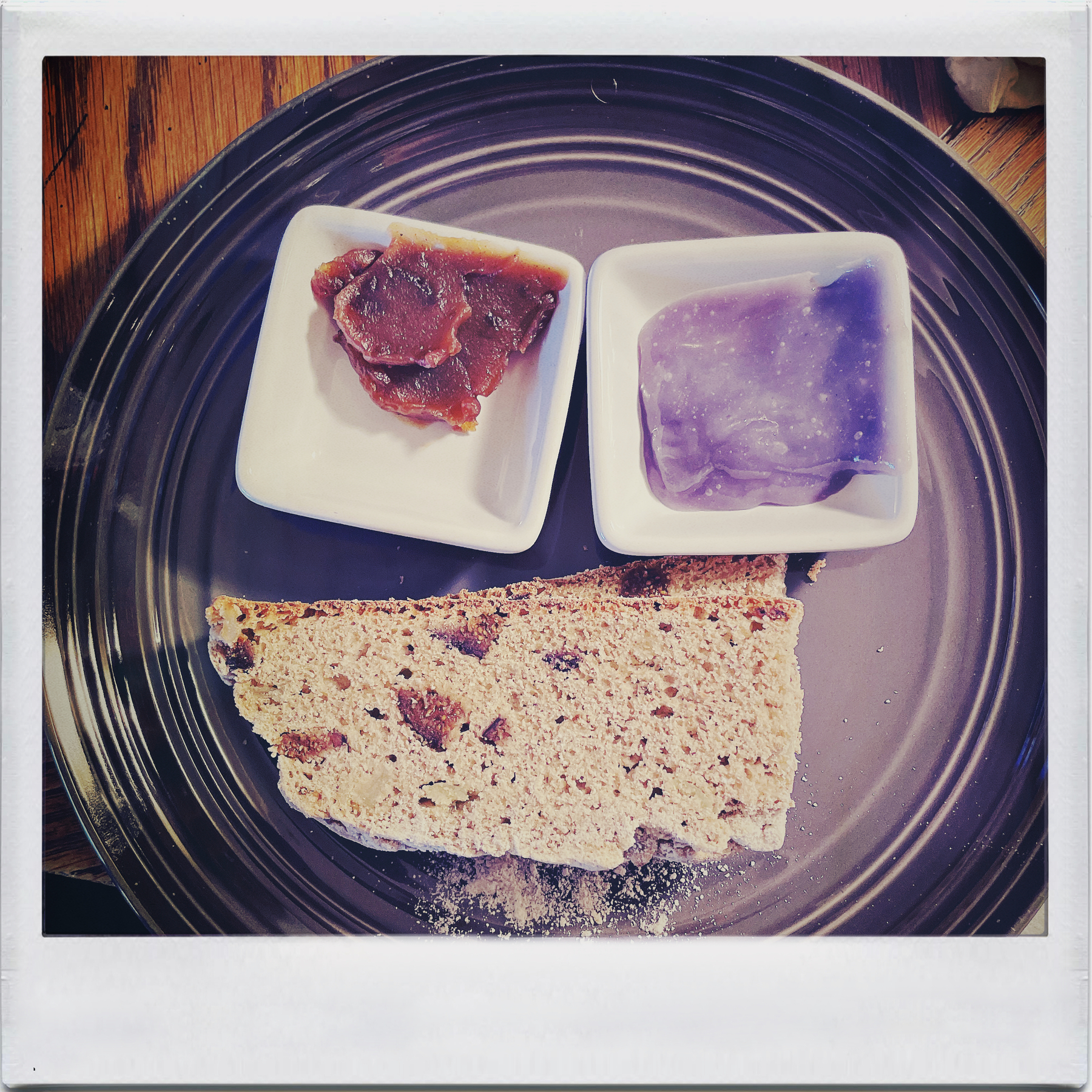 top-down view of a grey plate with a piece of fig and caramelized onion soda bread and two white square containers with red and purple spreads