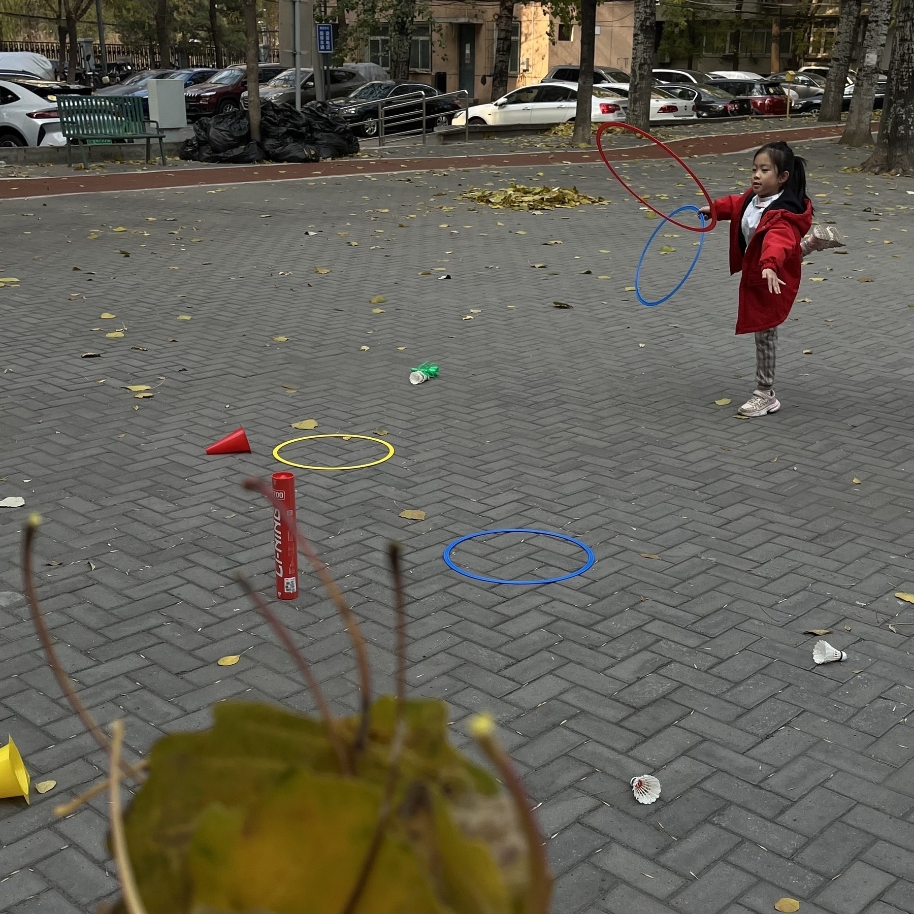 A child in a red coat plays with hula hoops on a paved area surrounded by fallen leaves.