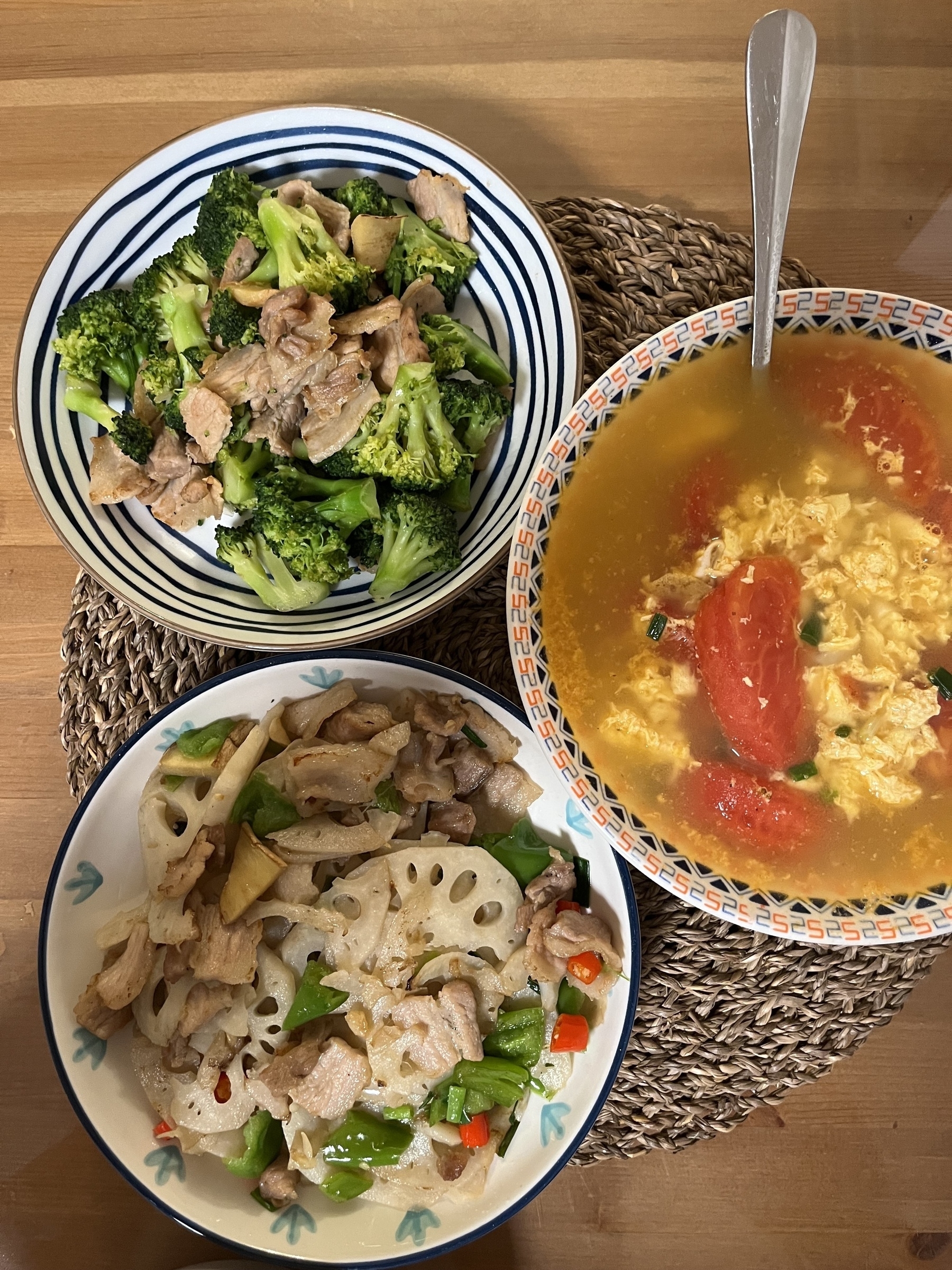 A meal featuring a bowl of scrambled egg and tomato soup, a dish of broccoli with meat, and another dish of lotus root and meat stir-fry all served on a woven placemat.