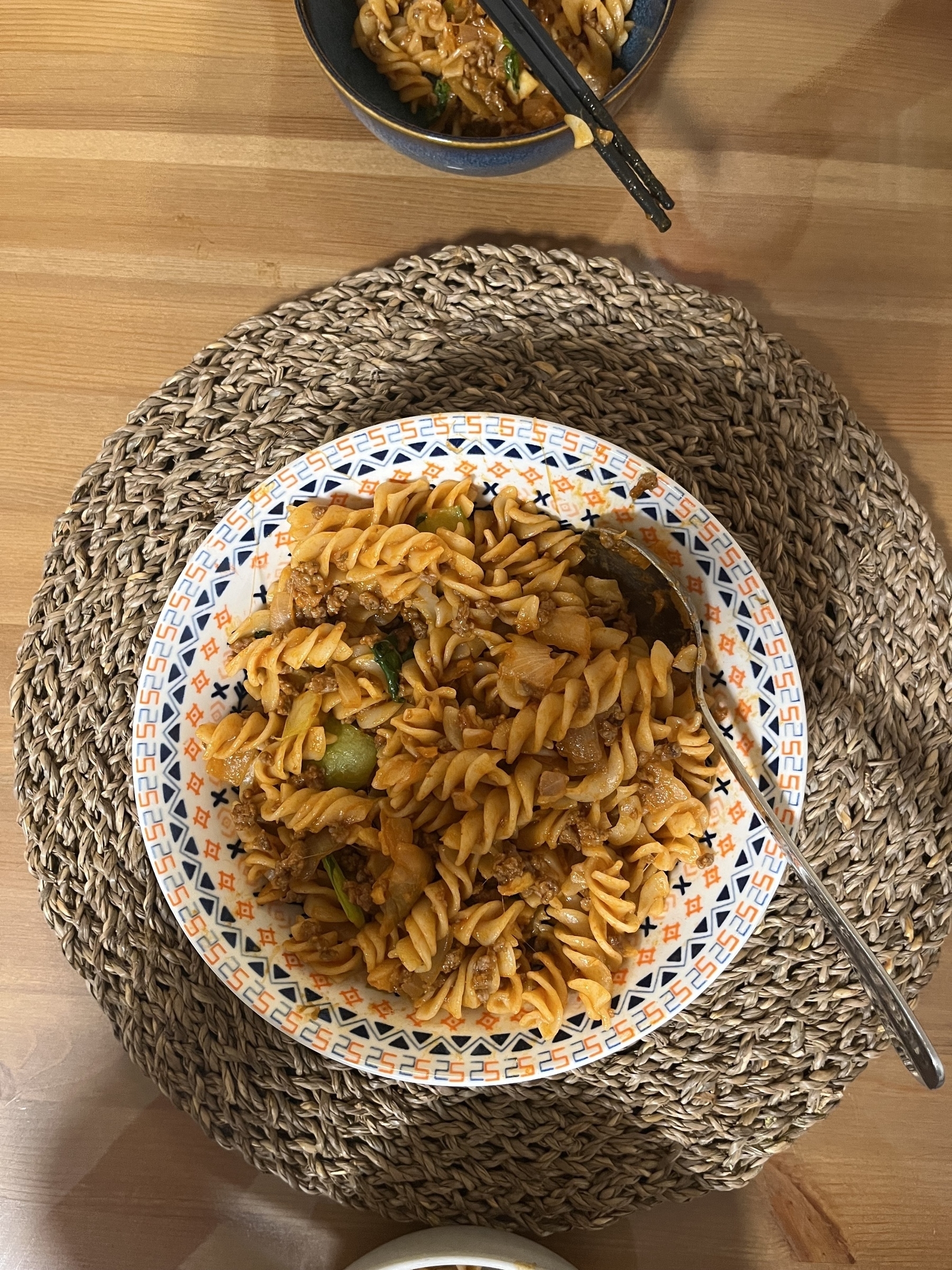 A plate of fusilli pasta with sauce and vegetables is served with a spoon on a woven placemat, accompanied by a small bowl of pasta.