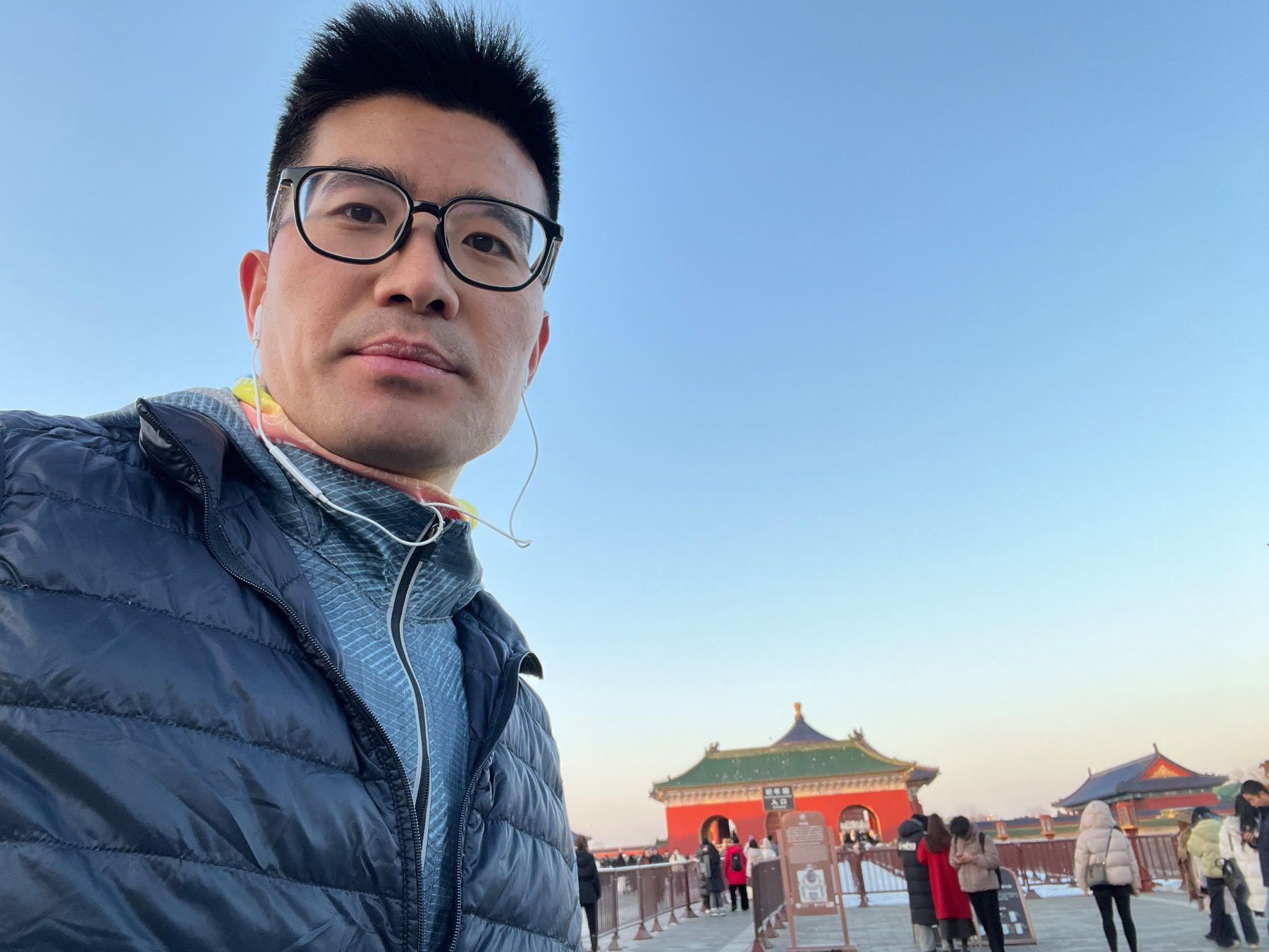 A person wearing glasses and a jacket takes a selfie with a traditional building and several people in the background.