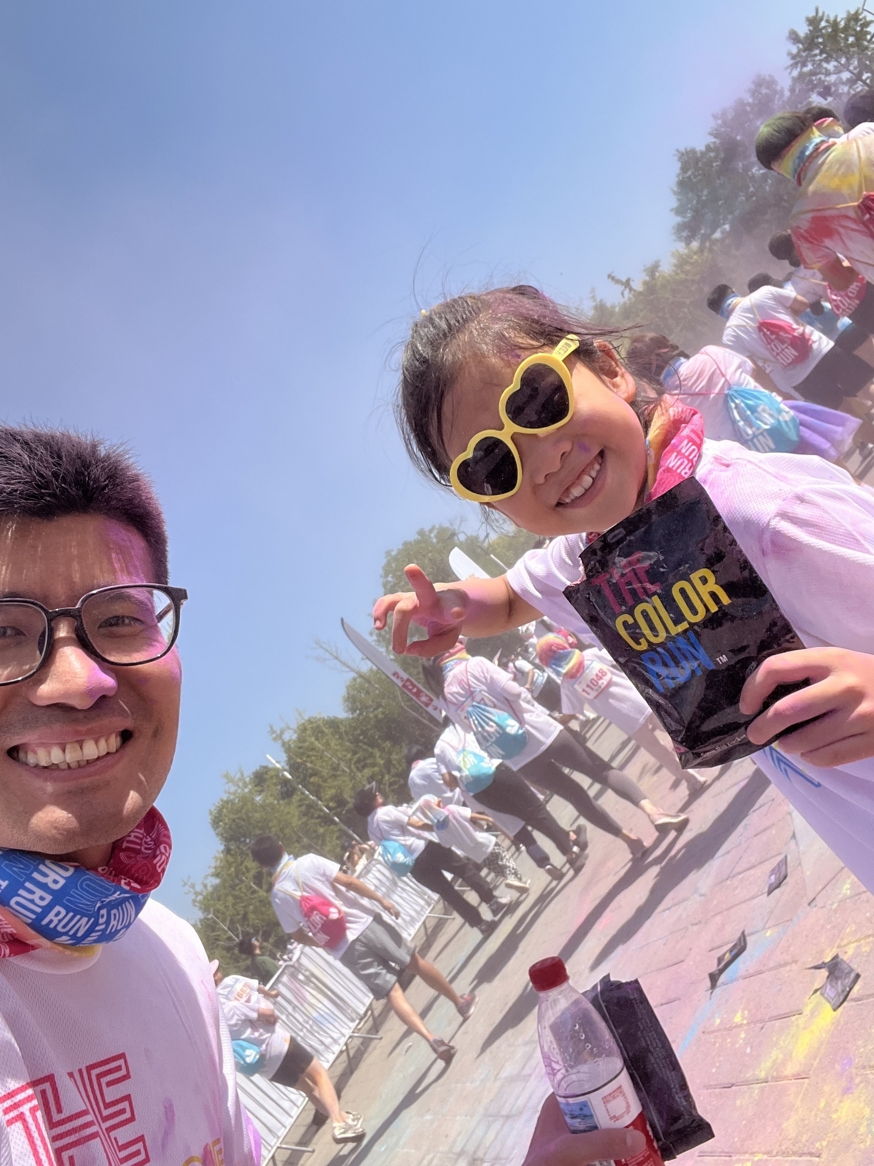 People are participating in a colorful outdoor event wearing white clothes with colorful powder, and a child is holding a packet of color while wearing heart-shaped sunglasses.