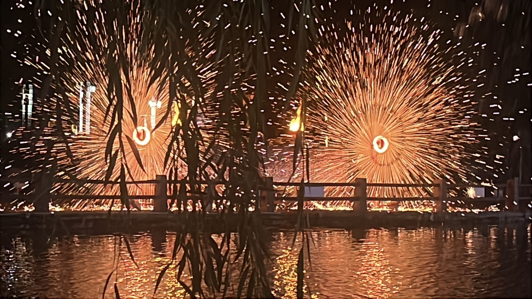 Two fiery displays resembling spinning wheels of sparks are seen against a backdrop, reflected in a body of water.