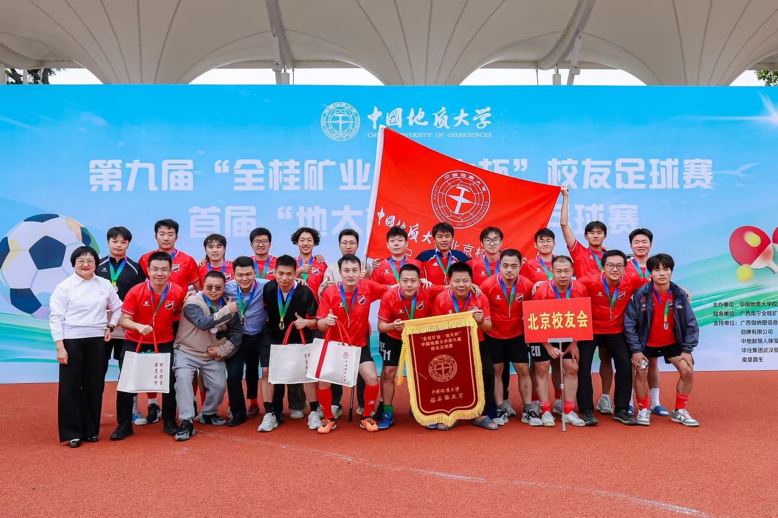 A group of soccer players wearing red uniforms pose with medals and a flag in front of a banner.