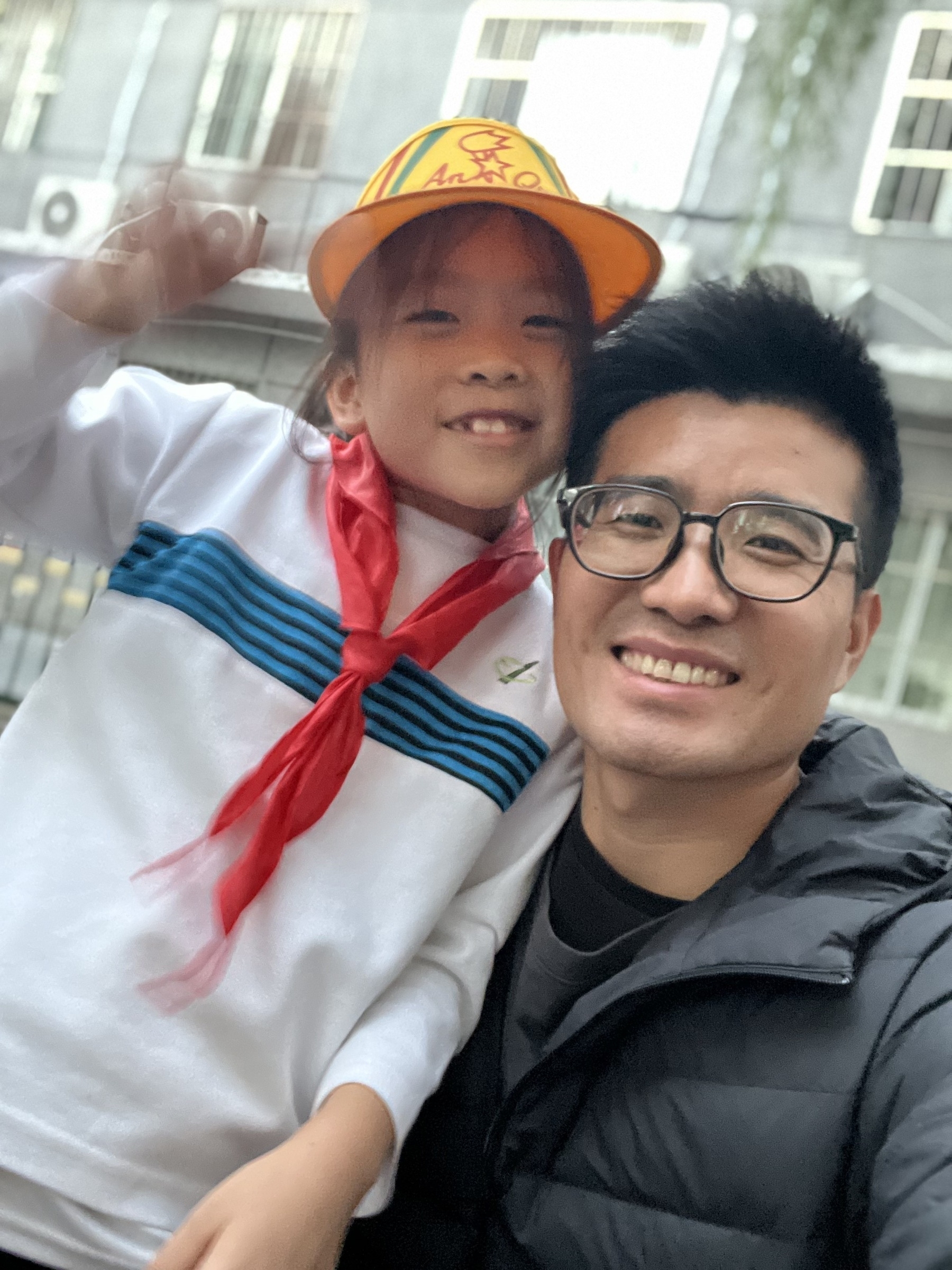 A man and a young child are smiling together, with the child wearing a yellow hat and red scarf.