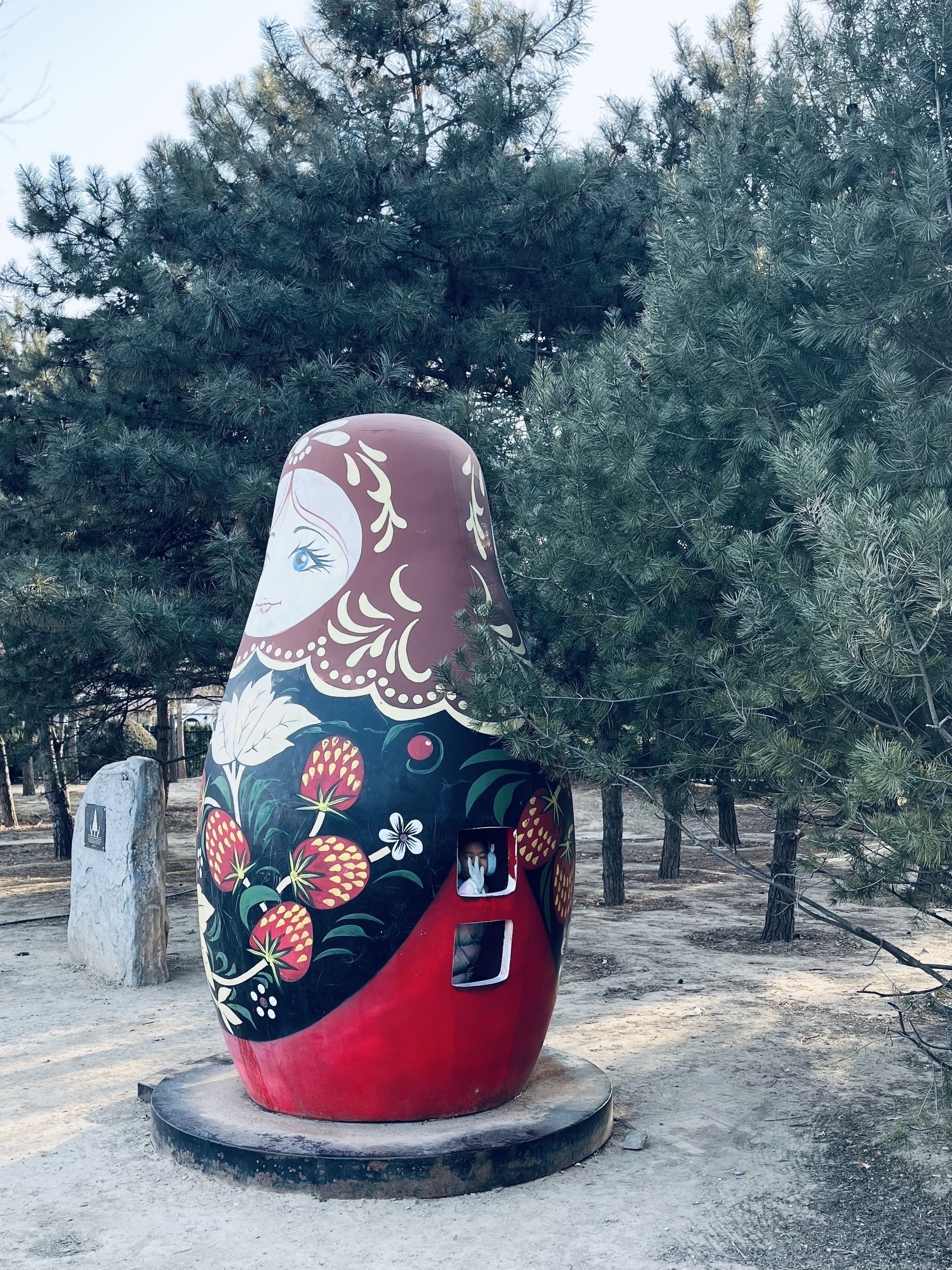 A large, beautifully painted Matryoshka doll sculpture is surrounded by pine trees in an outdoor setting.