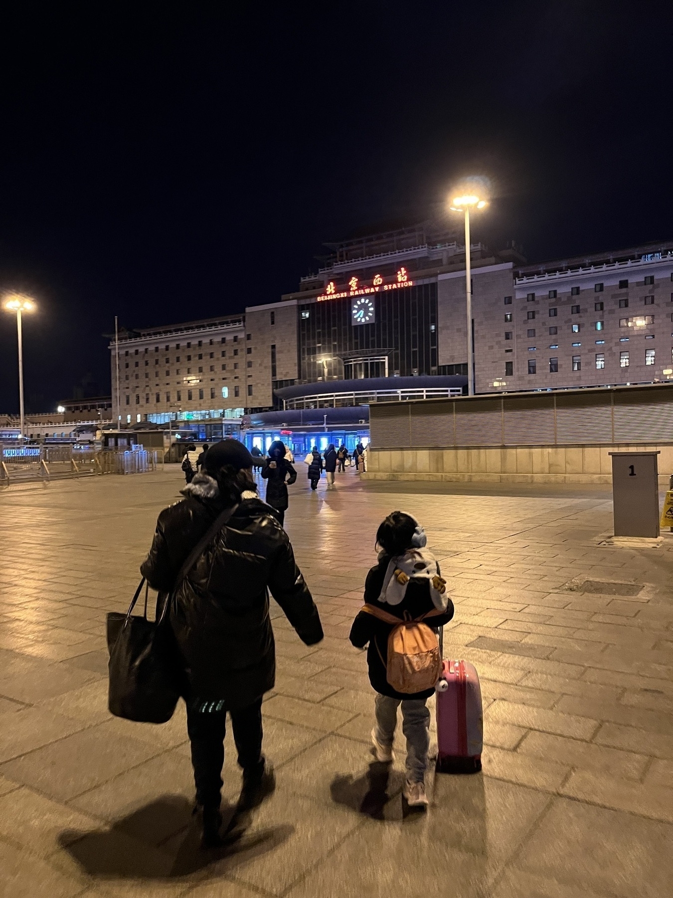 A person and a child with luggage walk towards a large, illuminated building at night.